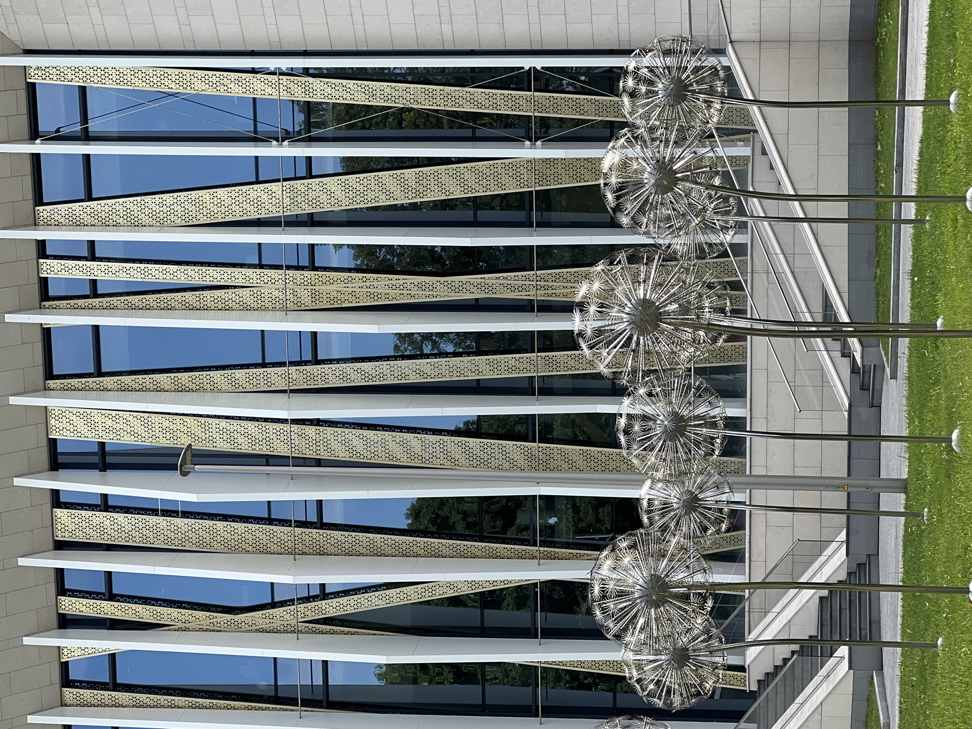 A modern building with a glass facade featuring a decorative metal screen stands behind a row of metallic dandelion sculptures planted in green grass. The building's facade is composed of vertical white and gold-colored panels arranged in a staggered pattern, interspersed with large blue glass windows. The sculptures, resembling oversized dandelions with their feathery seed heads, are made of polished metal and stand on thin poles. In the background, a set of concrete stairs with metal railings leads up to the building. A light fixture on a pole is visible near the center of the image. The overall scene suggests an outdoor public space or plaza in front of a contemporary architectural structure.