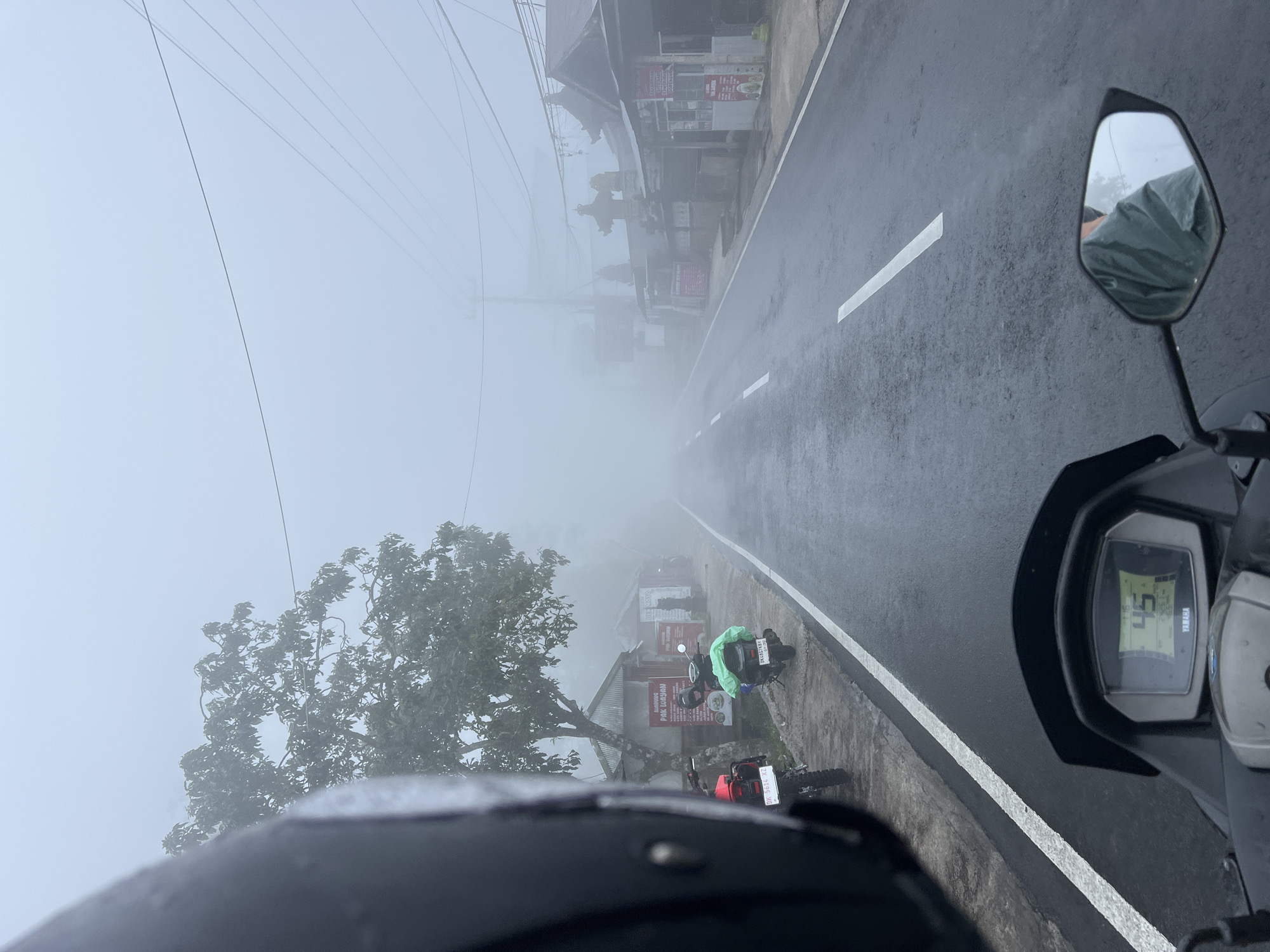 The image depicts a first-person perspective from a motorcycle traveling on a wet, two-lane asphalt road in Wanagiri, Indonesia, under conditions of dense fog. In the immediate foreground, the left side of a dark motorcycle helmet occupies the upper-left quadrant, exhibiting visible water droplets. Below, the dashboard of a Yamaha motorcycle is partially visible, with a digital display showing "45" km/h. The right rearview mirror reflects a portion of a person wearing a green garment.
Along the left side of the road, a concrete sidewalk is present. Adjacent to this sidewalk stands a large, leafy tree. Behind the tree, two modest structures are visible; one is identified by a sign reading "WARUNG PAK WAYAN," featuring a corrugated metal roof. Two motorcycles are parked on the sidewalk: a red dirt bike with license plate "DK 5616 XZ" and a darker scooter with a green covering on its rear, bearing license plate "DK 6467 KBV."
To the right of the road, a series of buildings display traditional Balinese architectural features, including ornate tiered roofs resembling small temples or shrines. Several signs with Indonesian text are affixed to these structures. Overhead power lines are visible spanning across the upper portion of the scene. The heavy fog significantly reduces visibility, obscuring distant elements and resulting in a uniformly pale, overcast sky. The road surface is visibly wet. No explicit human figures or direct interactions are discernible, beyond the implicit presence of the motorcycle operator.