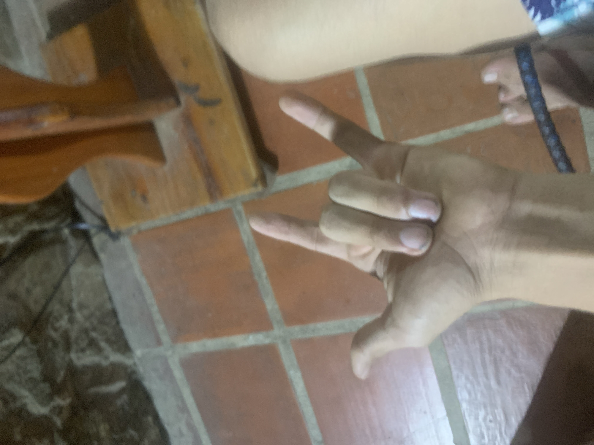 The image shows a close-up of a person's right hand in a "rock on" gesture, with the thumb, index finger, and pinky finger extended. The hand is positioned over a tiled floor. To the right of the hand, a portion of a person's leg and foot is visible, wearing flip-flops. In the background, there is a wooden structure and a stone wall. The scene appears to be indoors, possibly in a residential setting in Pedro González, Venezuela.