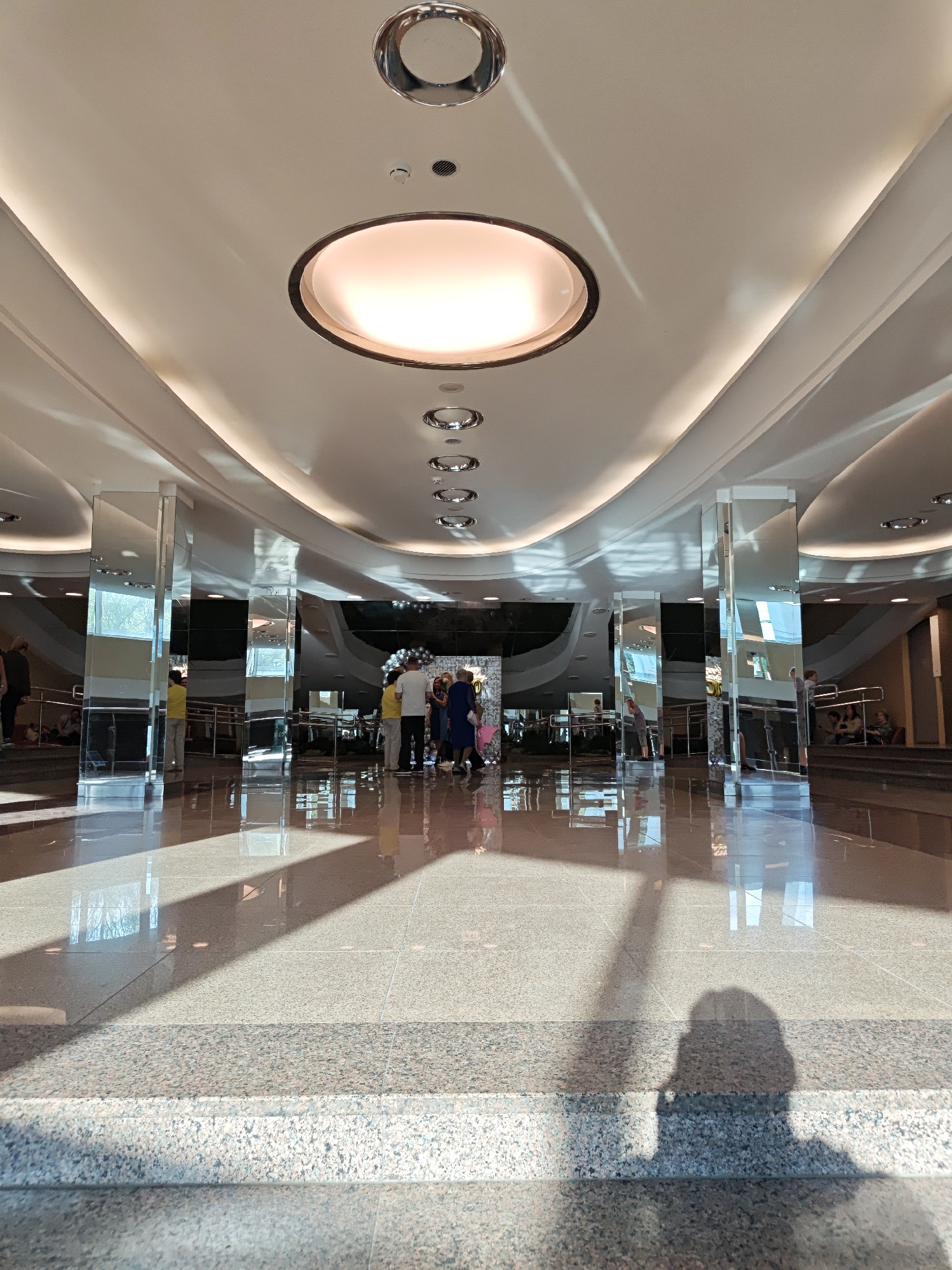 The image shows a spacious indoor lobby with a high, ornate ceiling featuring recessed lighting and decorative circular fixtures. The floor is polished granite, reflecting the light and the surrounding architecture. Several glass columns line the space, mirroring the scene and adding to the visual depth. In the background, a group of people is gathered near what appears to be a decorated area with balloons, suggesting a celebration or event. The location is likely the Hyatt Regency Yekaterinburg in Yekaterinburg, Russia, based on the distinctive architectural style and interior design elements. The lighting is bright, casting shadows across the floor and creating a sense of grandeur. In the foreground, a partial shadow of the photographer is visible.