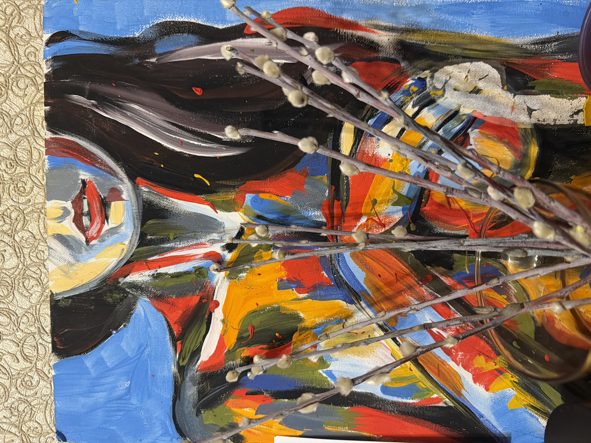 The image shows a close-up of an abstract painting with branches bearing furry buds placed in front of it. The painting features bold brushstrokes in a variety of colors, including blue, red, yellow, orange, black, and white. To the left of the painting, a section of a textured, cream-colored surface is visible. The branches with buds are the main foreground element, partially obscuring the painting. The details of any specific location in Naberezhnye Chelny, Russia, are not visible within the image.
