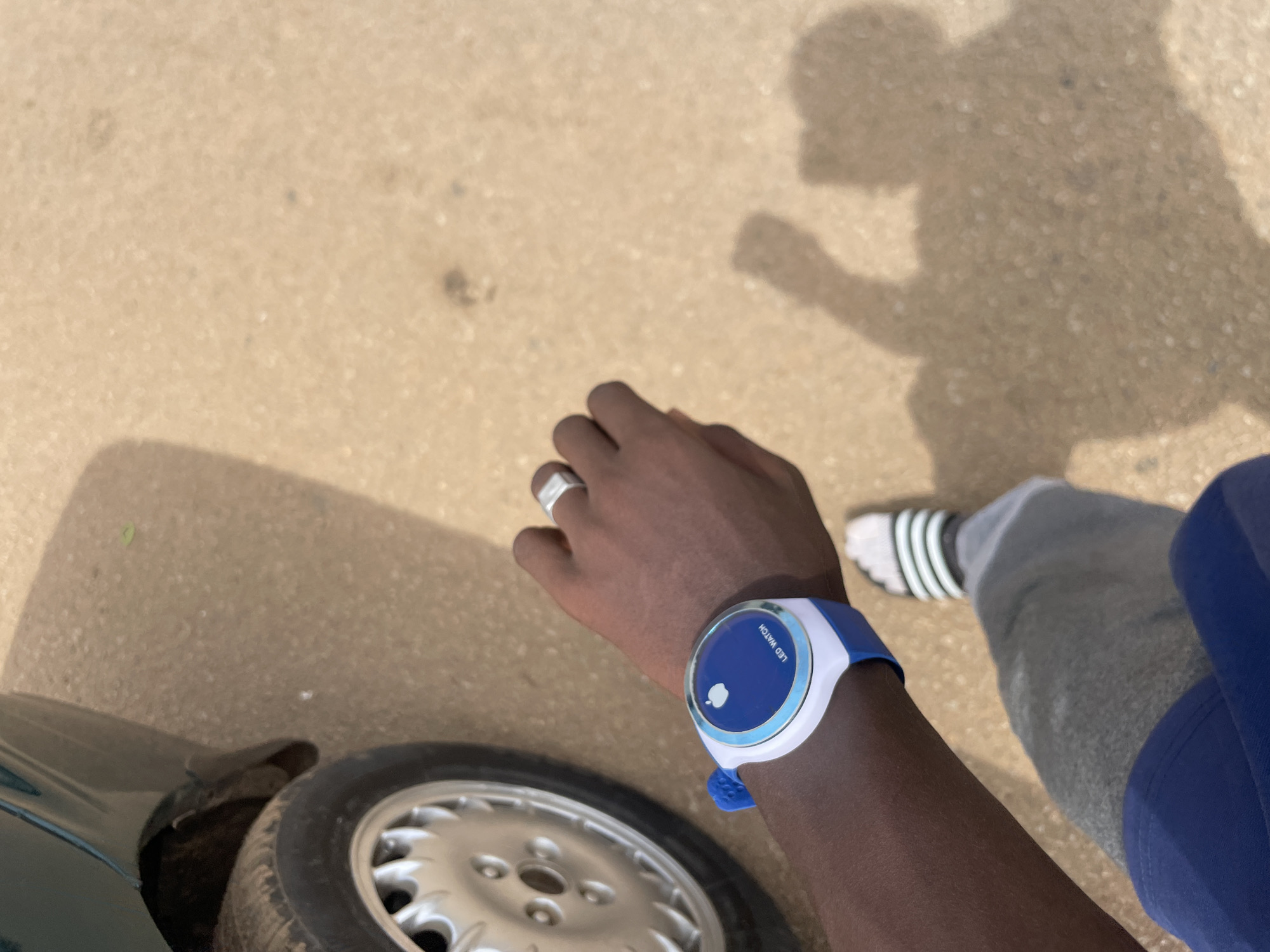 The image shows a close-up of a person's arm wearing a white and blue digital watch with an Apple logo and the words "LED WATCH" visible. The person is also wearing a silver ring on their right index finger. In the background, a car's tire and part of its body are visible, along with a sandy or gravelly surface that appears to be a road or pathway. The shadow of the person taking the photo is cast on the ground. The scene suggests an outdoor setting. It is not possible to determine the specific location within Jos, Nigeria from the provided image.
