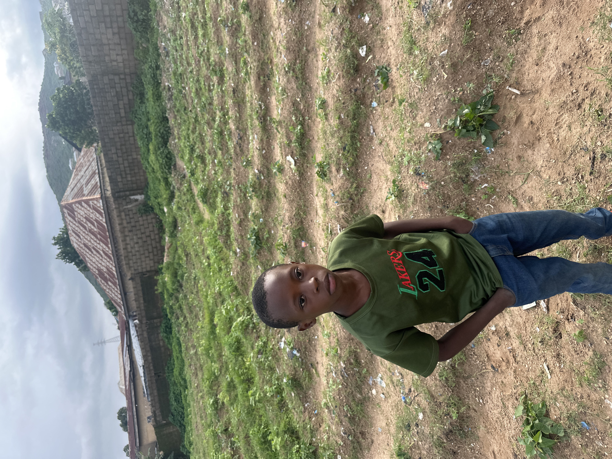 The image captures a young boy standing in an outdoor environment on Rukuba Road, Nigeria. The boy, appearing to be of African descent, is wearing a green t-shirt with "LAKERS 24" emblazoned on it and blue jeans. He is positioned in the foreground, with his hands in his pockets, and looking towards the camera with a neutral expression.
The background reveals a rural or semi-urban landscape. The ground is a mixture of dirt and sparse vegetation, with some visible debris. In the middle ground, there are rows of cultivated plants, suggesting some form of agriculture or gardening. Beyond the cultivated area, a brick wall separates the scene from further structures. These structures include buildings with corrugated metal roofs, some appearing to be residential. Further in the distance, a hill or mountainside is visible, partially obscured by greenery and cloudy skies. A tall transmission tower can also be seen in the far distance. The overall impression is of a somewhat neglected or developing area.