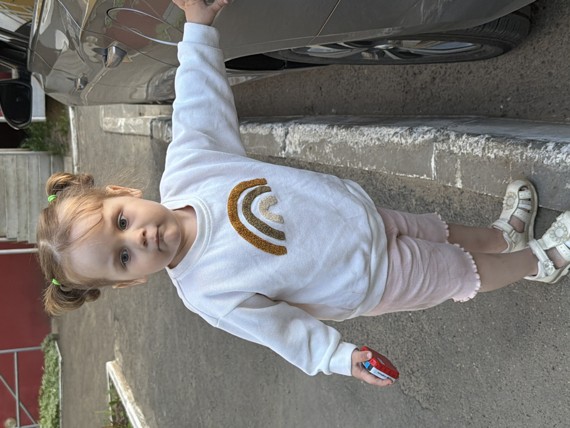 A young girl stands on a city street in Chaikovsky, Russia, next to a gray car. She wears a white sweatshirt with an embroidered rainbow, light pink shorts, and white sandals. She is holding a small rectangular object in her left hand. The scene also includes a curb and a building with a red exterior wall in the background.