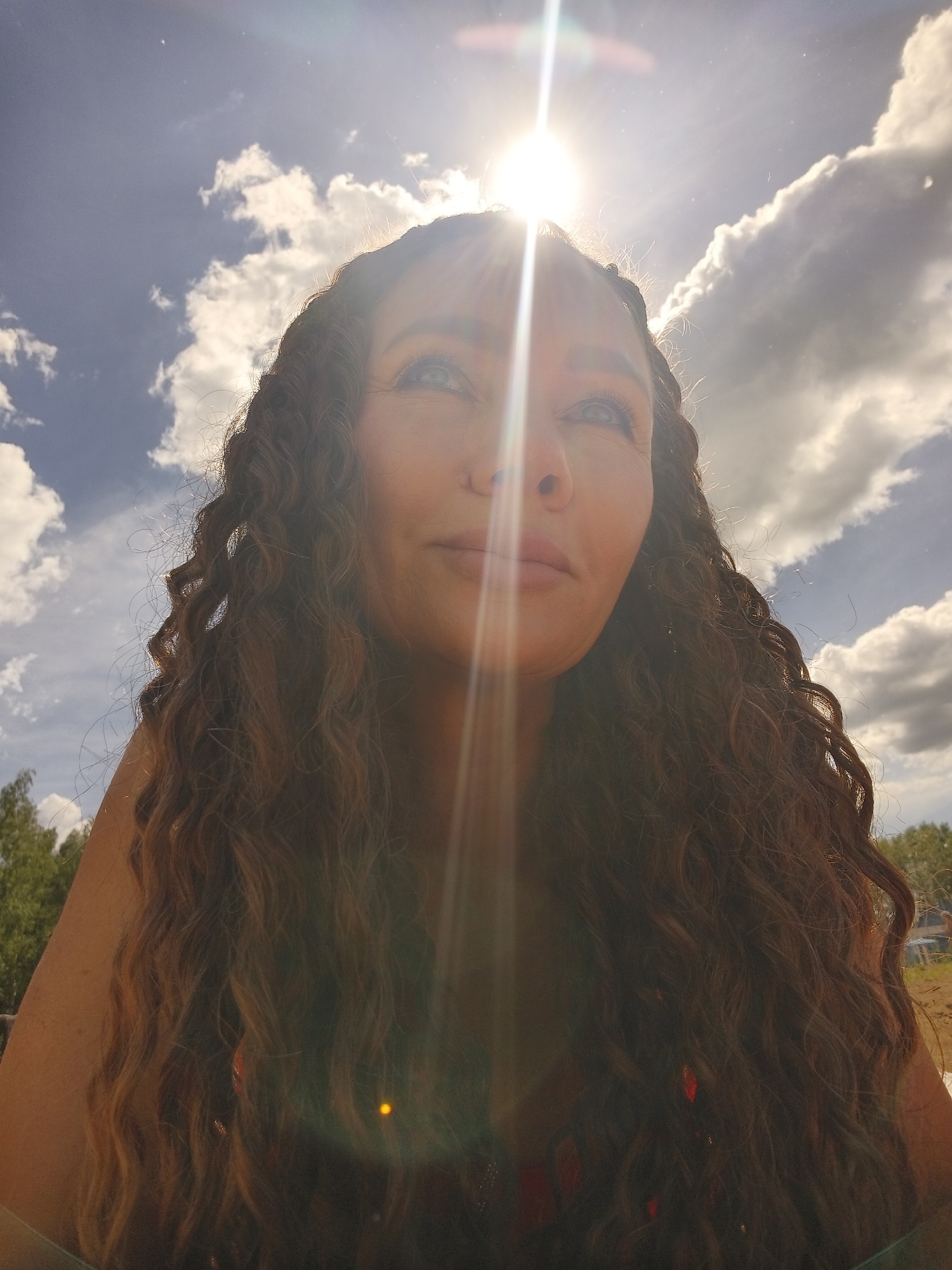 A close-up, low-angle shot shows a woman with long, dark, curly hair. She is looking up towards the sun, which is directly overhead and creating a lens flare effect that partially obscures her face and the sky. The sky is blue with scattered white clouds. The woman's hair is the most prominent feature, framing her face and extending down her shoulders. The sun's rays create bright streaks of light across the image, with some red and yellow lens flares visible. In the background, blurred trees and a hint of what appears to be a body of water or open land can be seen. The overall impression is one of being outdoors on a sunny day.