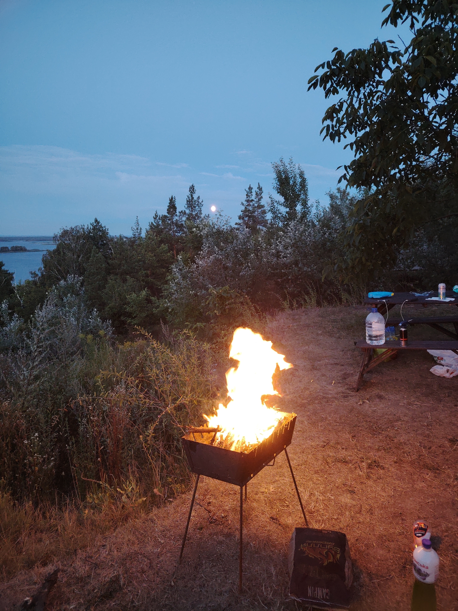 The image depicts an outdoor scene at dusk, likely in the city of Staiky, Ukraine. In the foreground, a portable charcoal grill with a bright fire is set up on dry grass. To the right of the grill, a cardboard box with Cyrillic text that appears to read "CAMPING" is visible, along with two plastic bottles, one of which has a purple cap and a label with Cyrillic text. Further to the right and slightly in the background, a wooden picnic table is set up. On the table, there are two plastic bottles of water, one larger and one smaller, a blue object, and a can. Behind the table, a dense cluster of trees and bushes creates a natural backdrop. In the distance, a body of water is visible, bordered by more trees and foliage. The sky above is a pale blue, with a faint haze, and a small, pale moon is discernible in the upper center of the frame. The overall impression is a campsite or picnic area during a twilight gathering.
