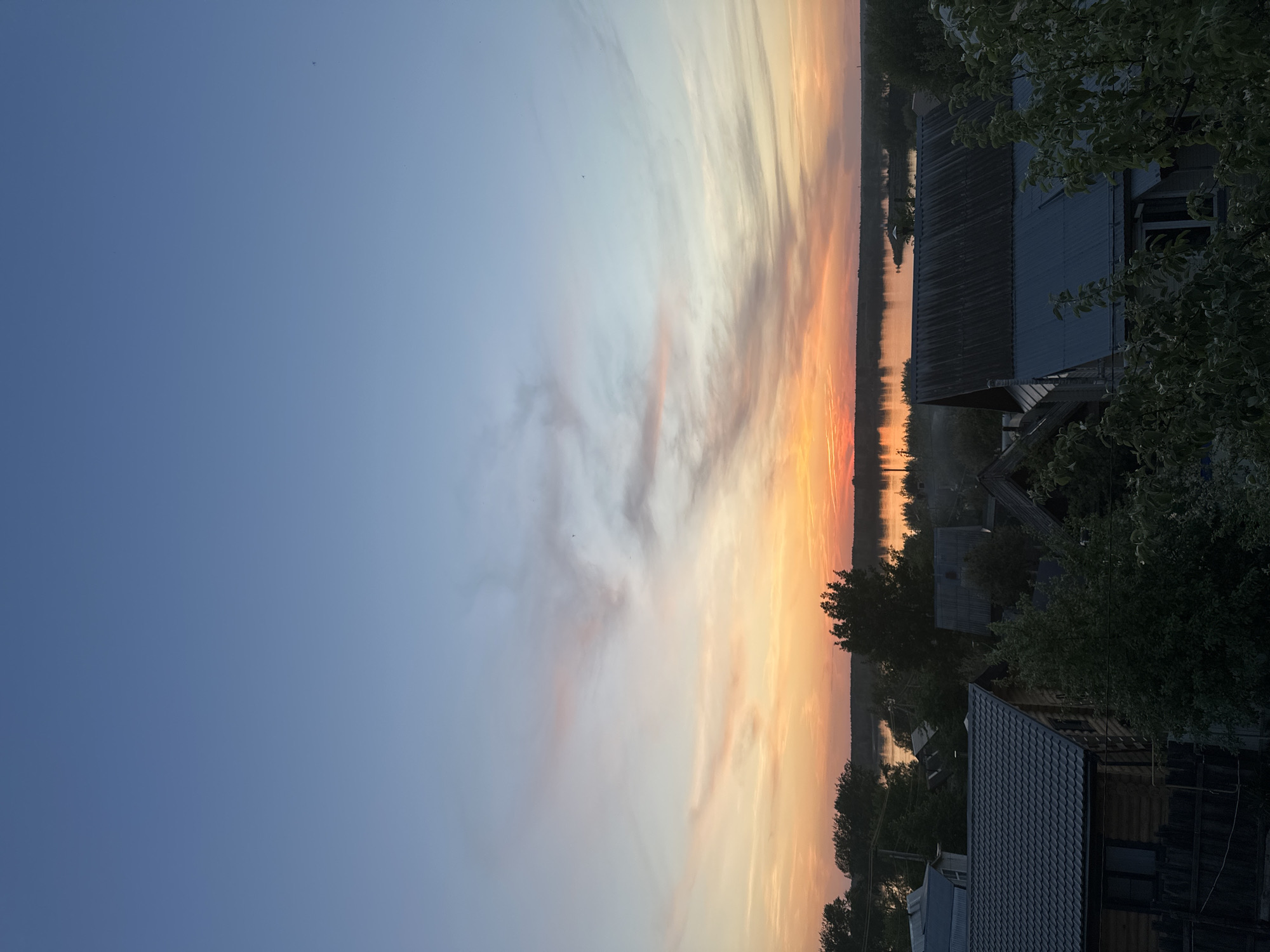 The image shows a landscape at dusk, possibly in Chaikovsky, Russia. The sky transitions from blue to orange, indicating a sunset. The land features buildings, trees, and a body of water, possibly a river or lake. The buildings are primarily residential, with dark roofs. Trees populate the areas surrounding the structures. The body of water reflects the light from the sky.