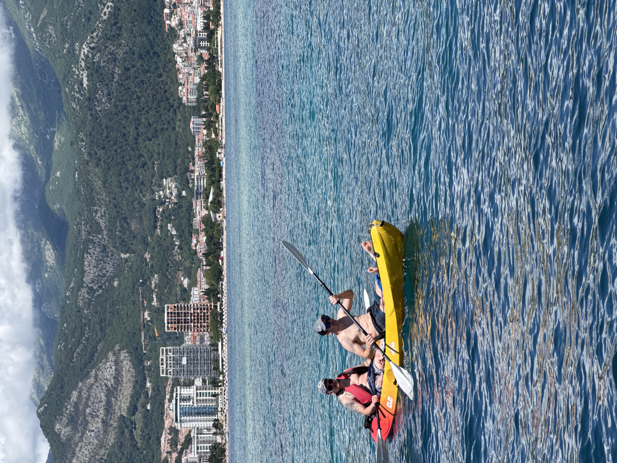 The image captures a vibrant coastal scene in Budva, Montenegro, under bright daylight. In the foreground and midground, two individuals are seen paddling kayaks on clear, blue-green waters that exhibit gentle ripples and strong sun reflections. One person, wearing a baseball cap and sunglasses, is in a yellow sit-on-top kayak, actively paddling with a double-bladed oar. Another person, also wearing a cap and sunglasses, and a red life vest, is visible in a red and orange kayak, with a paddle partially visible. On the side of this kayak, the text "OLD" is discernable.
The background features a densely developed coastline, characteristic of Budva, with numerous buildings ranging from traditional structures with red roofs to modern high-rises. These buildings stretch along the shore, backed by impressive, steep, and heavily forested mountains that rise dramatically. Some white clouds are visible clinging to the upper slopes and peaks of these mountains, adding depth to the landscape. The overall impression is one of active recreation in a beautiful, Mediterranean setting, with a clear sky and calm waters.