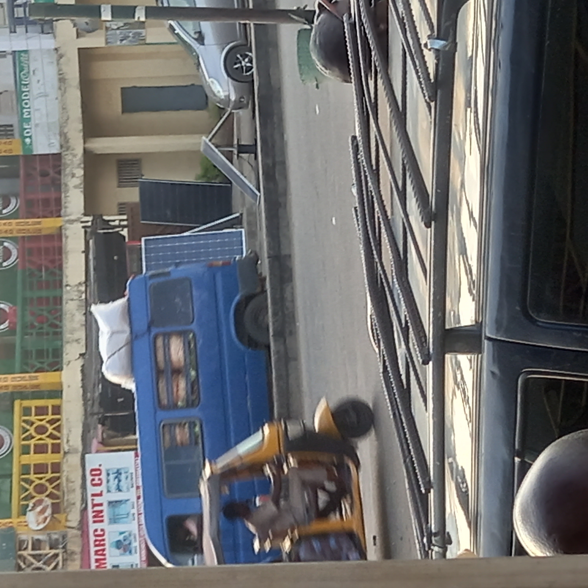 The image, taken in Dadi Riba, Nigeria, captures a vibrant street scene during daylight hours under clear weather conditions. The perspective appears to be from within a vehicle, looking across a road populated by various modes of transport.
On the left, a building facade is visible, featuring stacked colorful plastic crates, including red, green, and yellow ones. Partial text like "D C" and "KO HOUSE" can be seen on these crates. A green sign above reads "OF MODEL Outfit," while a red and white sign below clearly displays "MARC INT'L CO."
In the mid-ground, a blue mini-bus, possibly a public transport vehicle, is prominent. White sacks are secured on its roof, and a small solar panel is visible towards its rear roof section. Another solar panel is positioned on the ground or a low structure behind the bus. Further down the street, a silver car is parked.
Closer to the viewer, a yellow motorized tricycle, commonly known as a Keke Napep, is visible with a person driving it. The right side of the frame is occupied by the dark structure and windows of the vehicle from which the photograph was taken, suggesting a protective grille or bars. The scene conveys typical daily street activity in an urban or semi-urban setting.