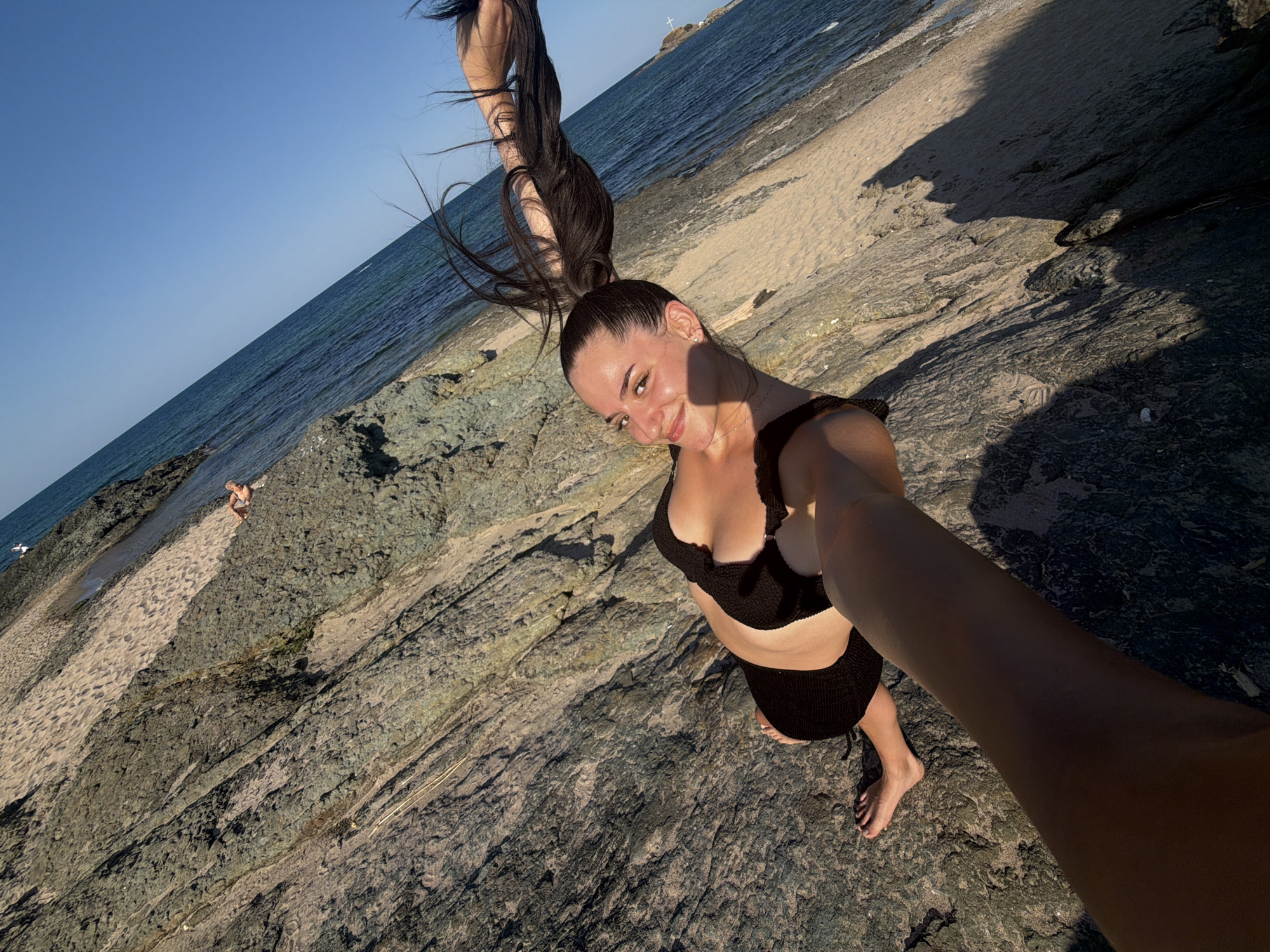 In Lozenets, Bulgaria, a young woman with long, dark hair pulled into a high ponytail poses for a selfie on a rocky beach. She is wearing a black bikini. Her hair is styled to extend outwards, creating a dynamic visual effect. The background shows the clear blue sea and a sandy shoreline. The image is taken from a low angle, with the photographer's arm extending into the frame. The lighting suggests it is late afternoon or early evening, casting shadows on the rocks. A second person is visible in the distance, further down the beach.