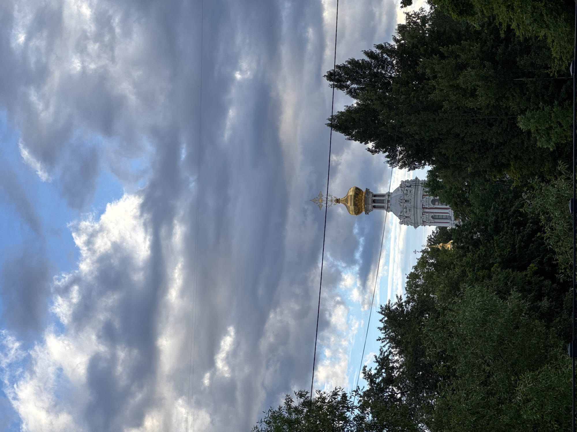 The image depicts the bell tower of the Smolny Cathedral in Saint Petersburg, Russia, seen through a canopy of lush green trees. The bell tower, with its white facade and ornate red and gold detailing, is topped by a golden dome and a cross. The sky above is dramatic, filled with a mixture of dark, heavy clouds and patches of bright blue, suggesting a transition in weather. Power lines are visible across the sky, partially obscuring the view. The foreground is dominated by the dense foliage of trees, with a dark fence or railing visible at the very bottom of the frame. The overall impression is one of a serene, architectural landmark partially revealed by nature, under a dynamic sky.