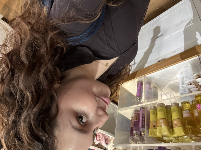 A woman with curly brown hair and a dark top stands next to a shelf stocked with Bath & Body Works products. The shelf has various lotions, perfumes, and sanitizers. The woman's face is turned towards the camera, and her eye is visible. There appears to be a shadow cast behind the shelf. The environment suggests the interior of a Bath & Body Works store.