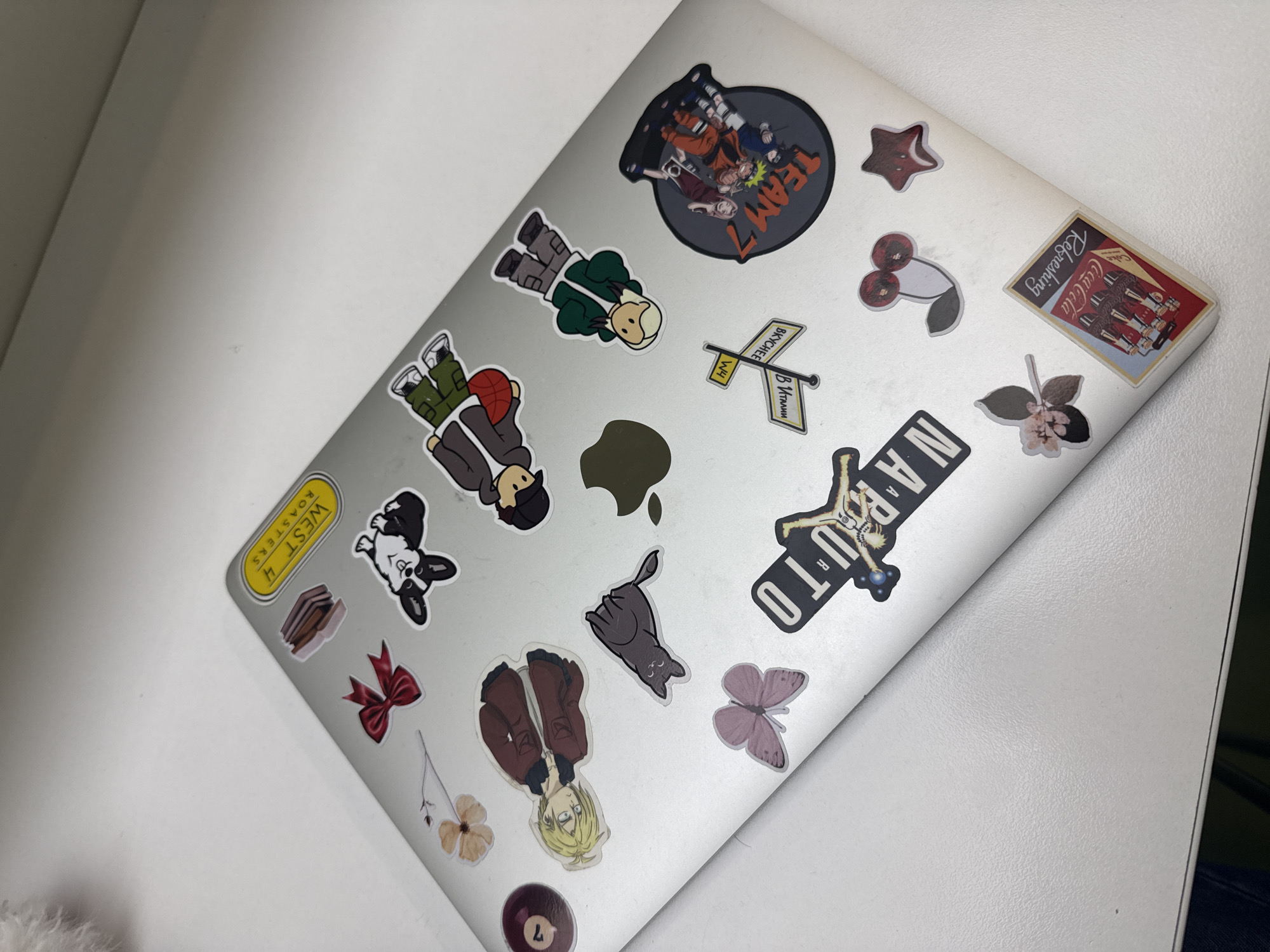 The image shows a laptop covered with various stickers. Among the stickers are images related to the anime Naruto, including the "Team 7" sticker and a stylized "NARUTO" text sticker. There's also a sticker with a Coca-Cola advertisement, a "WEST 4 ROASTERS" sticker, a sticker with the words "В ИТАЛИИ BKYCHEE," and several other stickers depicting anime characters, animals, and miscellaneous objects. The laptop rests on a white surface. There is no indication about the city of Yerevan, Armenia.