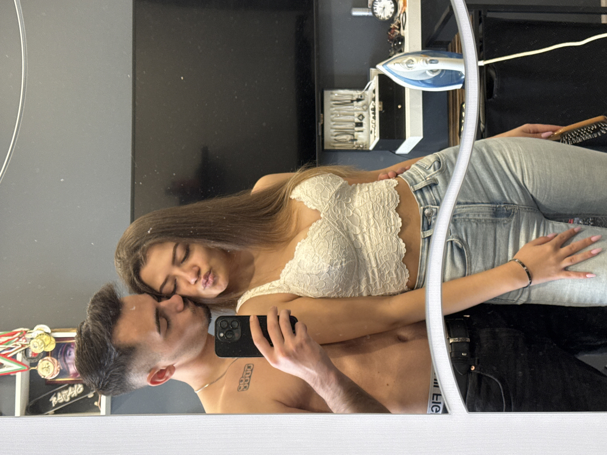 A couple is reflected in a mirror. The woman is wearing a white top and light blue jeans. The man is shirtless and holding a black smartphone. They are posed close together, as if taking a selfie. Behind them in the reflection, there is a TV, a grooming kit, an iron, and some shelves. The location, Sofia, Bulgaria, cannot be determined from the information in the image.