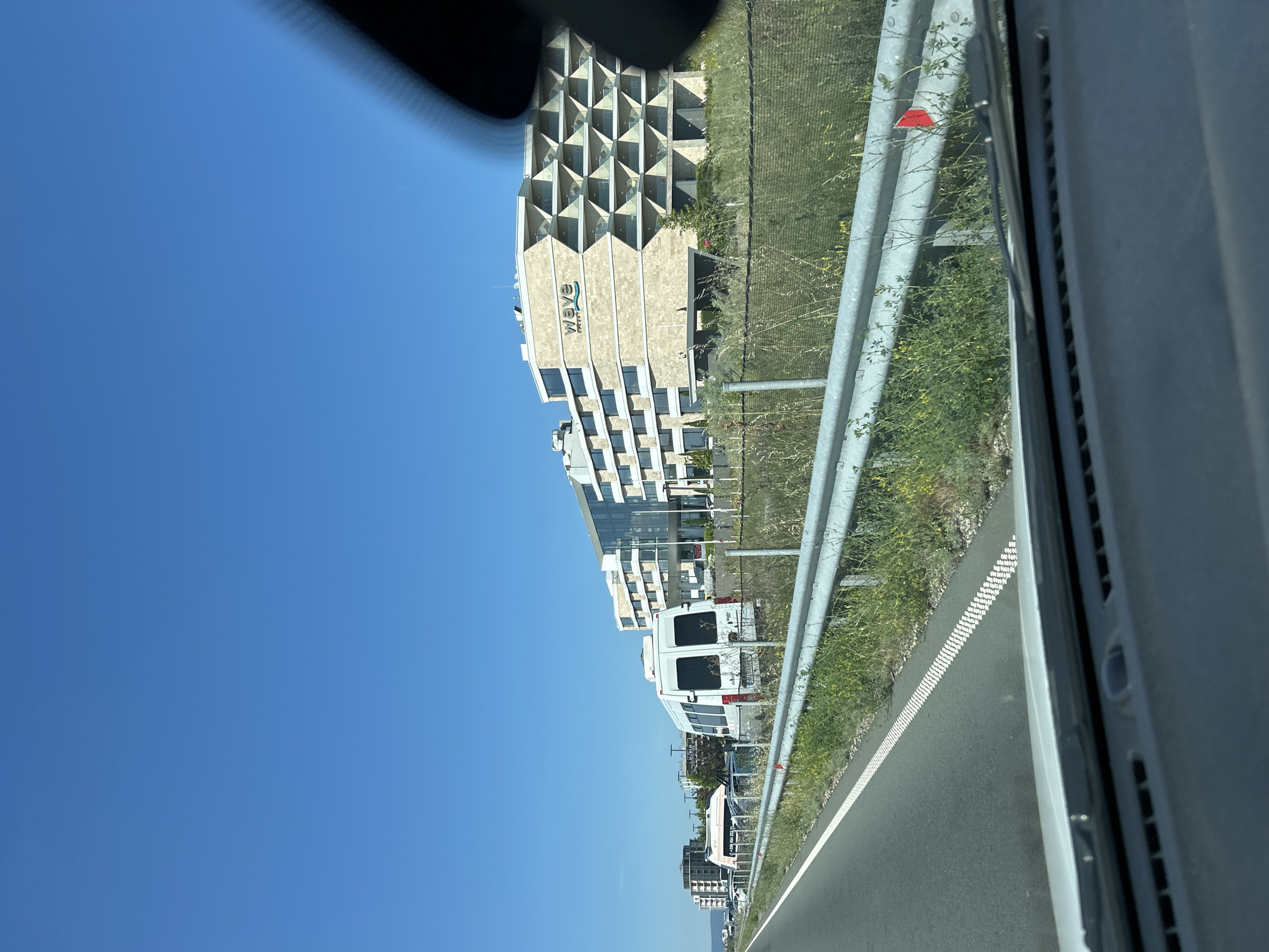In Aheloy, Bulgaria, a highway scene unfolds under a clear blue sky. On the right, a modern, multi-story hotel with a distinctive honeycomb-like facade is visible, branded with "wave" and "hotel" in its logo. The hotel is set back from the road, separated by a grassy area and a metal fence. To the left of the highway, a white van is parked on the side of the road, with another vehicle partially visible behind it. The road itself is asphalt with white lane markings. The foreground shows the dashboard and windshield of a vehicle, suggesting the photo was taken from inside a car driving on the highway. A red marker flag is attached to the highway barrier in the middle ground. The overall environment is sunny and clear, with some dry grass and weeds growing along the roadside.