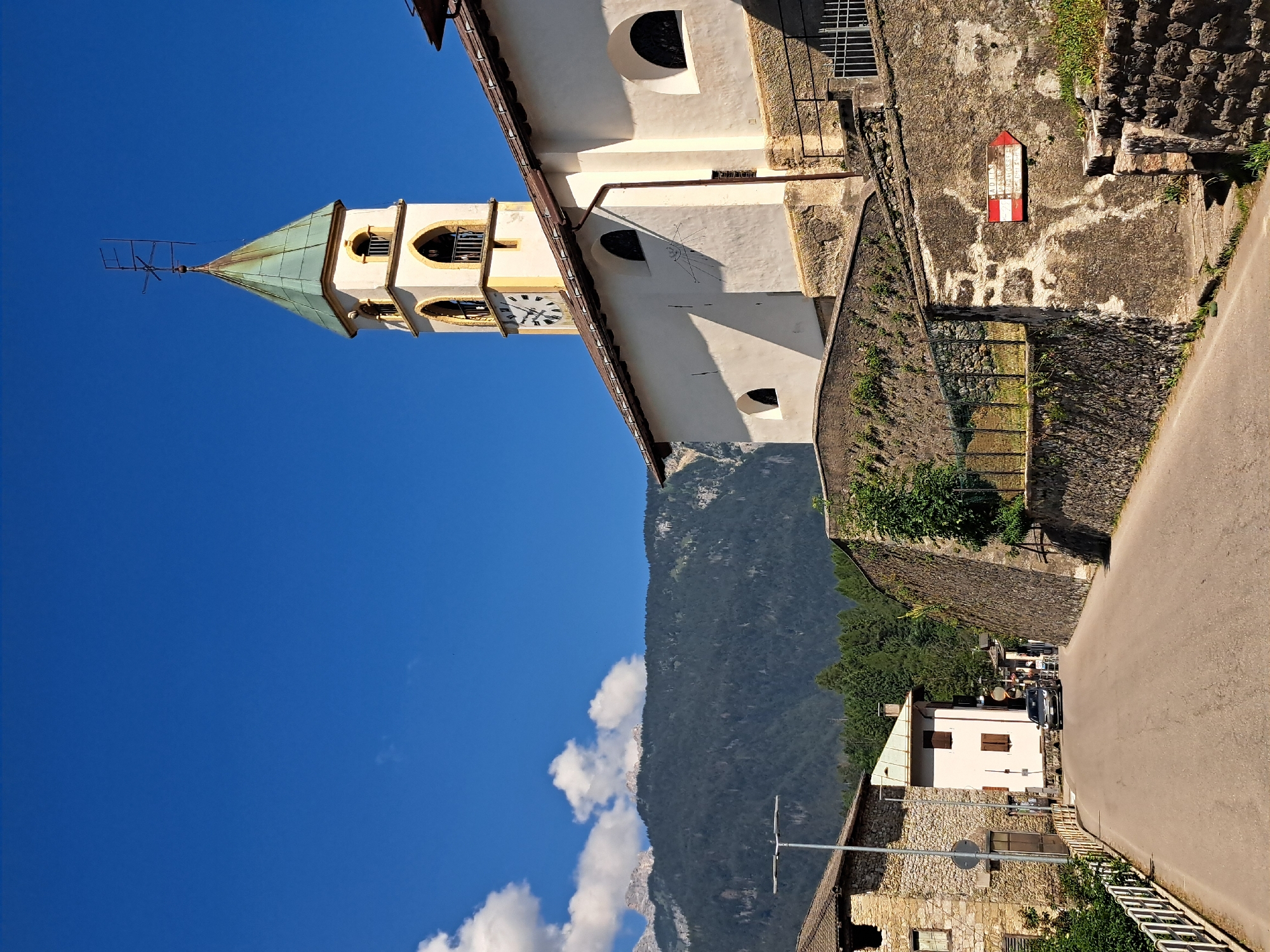 In a sunlit outdoor scene, a street curves uphill towards a village. On the right, a white church with a tall bell tower rises, its steeple capped with a green, aged copper roof. The church features arched windows and a sundial on its facade. A stone wall surrounds the church, adorned with a red and white directional sign pointing to the right. The street is paved and lined with stone buildings on the left, one with a rough stone exterior and a light-colored facade. A dark-colored car is parked on the street. In the background, a dense, dark green forest covers the slopes of a mountain range under a clear blue sky with scattered white clouds. The overall impression is a peaceful, rural setting in a mountainous region.