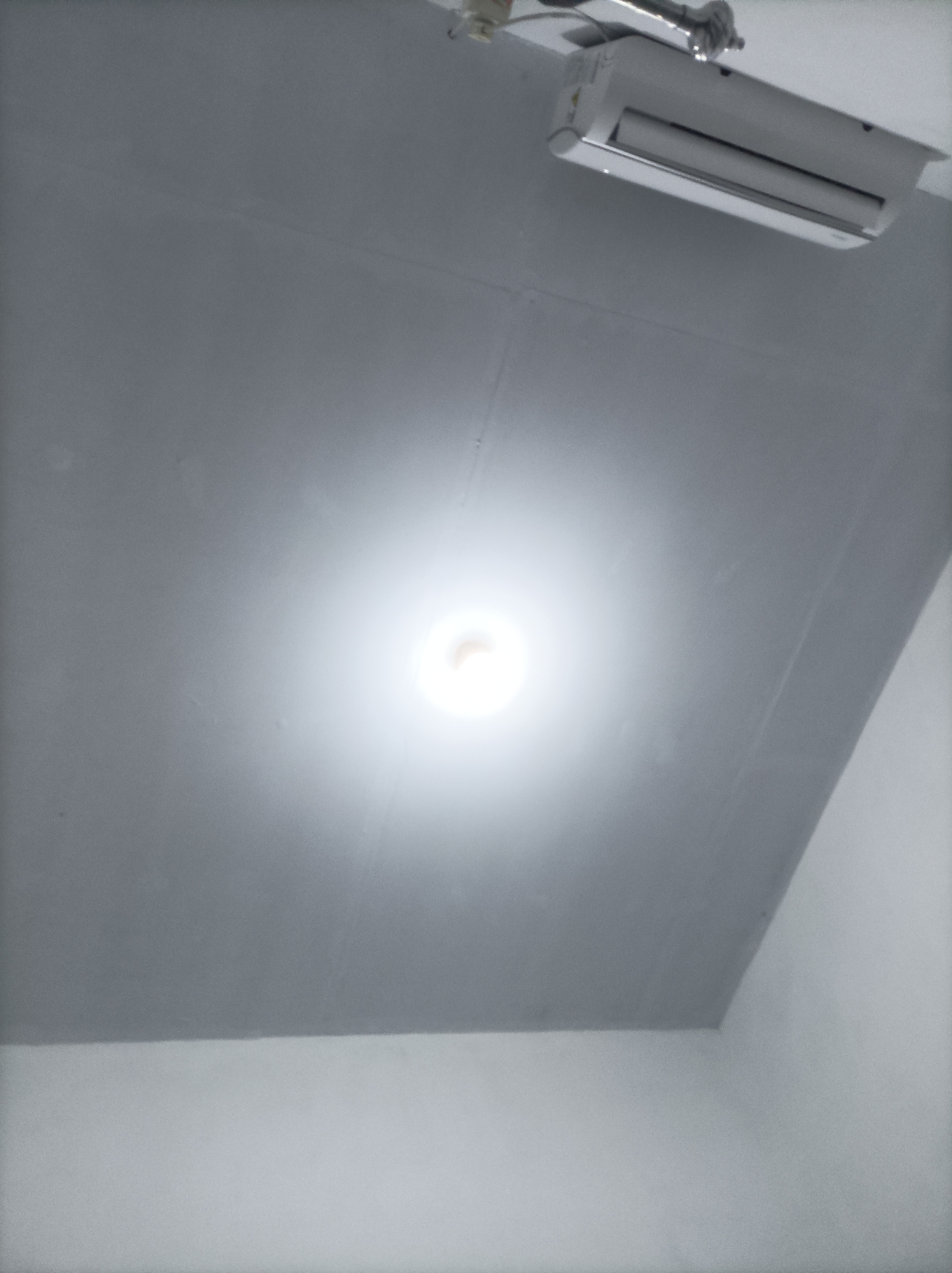 The image shows a white air conditioning unit mounted high on a grey ceiling. Below the air conditioner, a bright light emanates from a light fixture on the ceiling, creating a halo effect. The ceiling appears to be painted a matte grey and shows some subtle lines or seams. The overall environment suggests an indoor space, possibly a room in a residential or commercial building in Pamengkang, Indonesia. There are no people visible in the image.