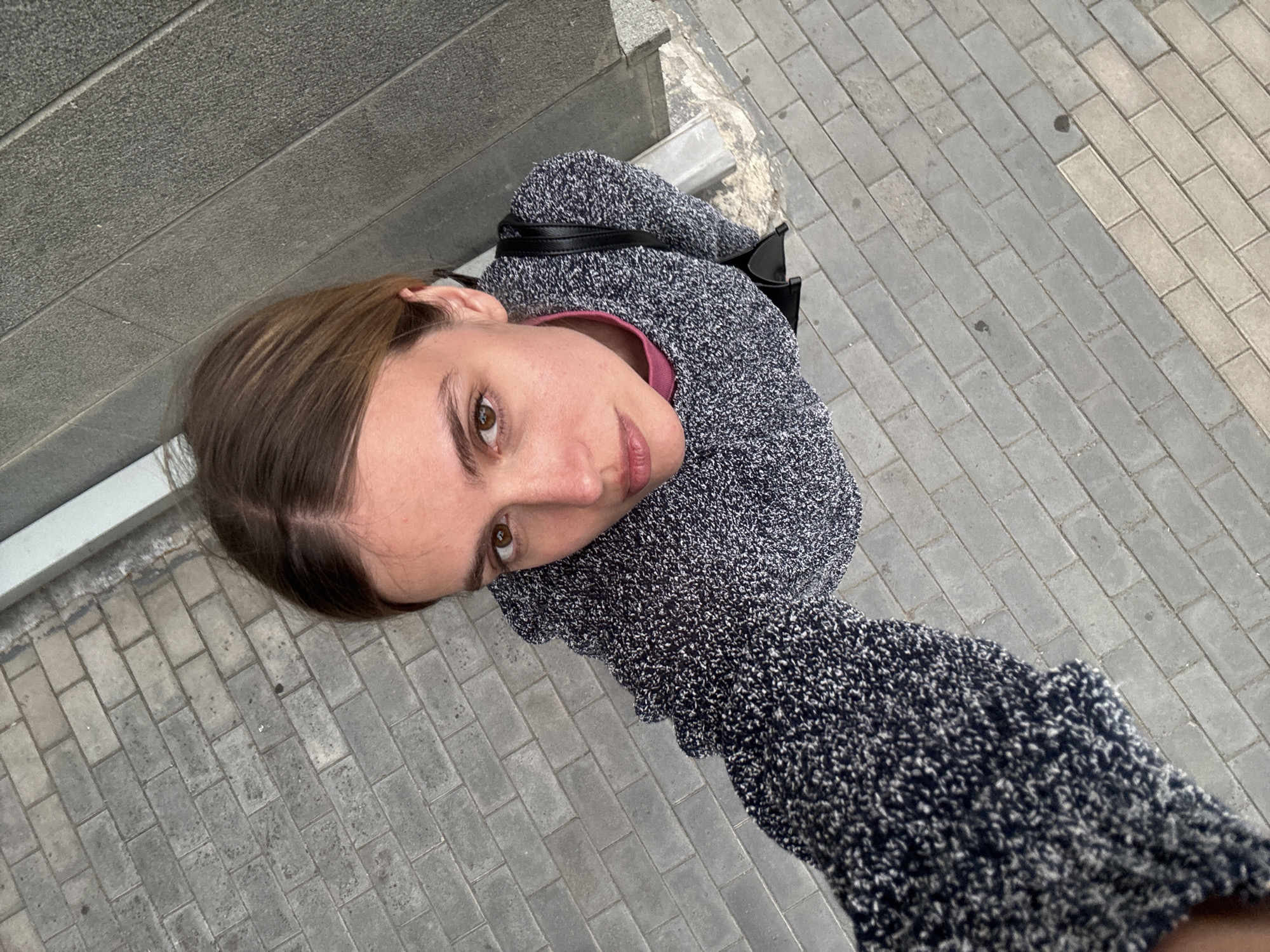 A woman with brown hair and a gray sweater is taking a selfie while standing on a gray brick walkway. The background includes a gray wall and part of the walkway. The location is in a city of Unknown, Unknown.