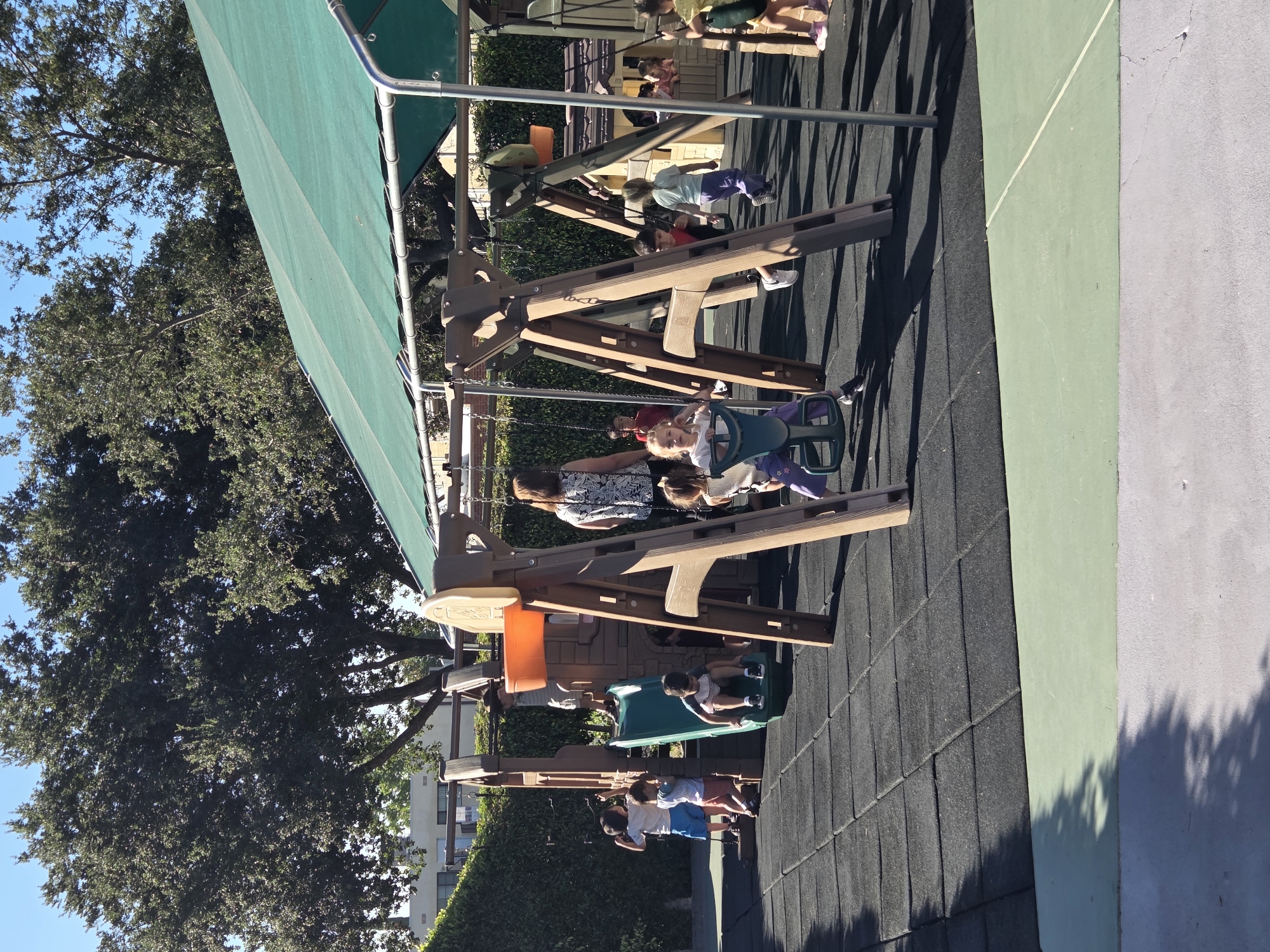 The image captures a vibrant outdoor playground scene on a sunny day, likely in Glendale, California, United States of America, given the presence of mature oak trees common in the region and the landscaping style. The foreground features a dark grey, rubberized playground surface composed of interlocking tiles, with a lighter green border along the bottom edge of the frame. A light grey concrete walkway with visible cracks runs parallel to the green border, casting dappled shadows.
Dominating the mid-ground is a large, multi-colored plastic and wooden playground structure. This structure includes a green slide, multiple platforms, and a swing set with two swings. Several children are actively engaged on the playground. One child is midway down the green slide, wearing a striped shirt and dark shorts. Another child, in a white shirt and blue shorts, stands at the base of the slide. A girl in a white top and purple pants is seated on a swing to the left, and another girl in a yellow top and blue shorts is on a swing to the right. In the background, more children are visible near a smaller playhouse structure, some peeking over its railing.
A woman with dark, curly hair, wearing a patterned black and white top, stands near the play structure, observing the children. To her left, a man in a light-colored hat and striped shirt stands on a platform of the play structure. Lush green hedges form a dense background to the left and behind the playground, with large, leafy trees reaching towards the clear blue sky. The overall impression is one of a lively and safe environment for children's recreation.