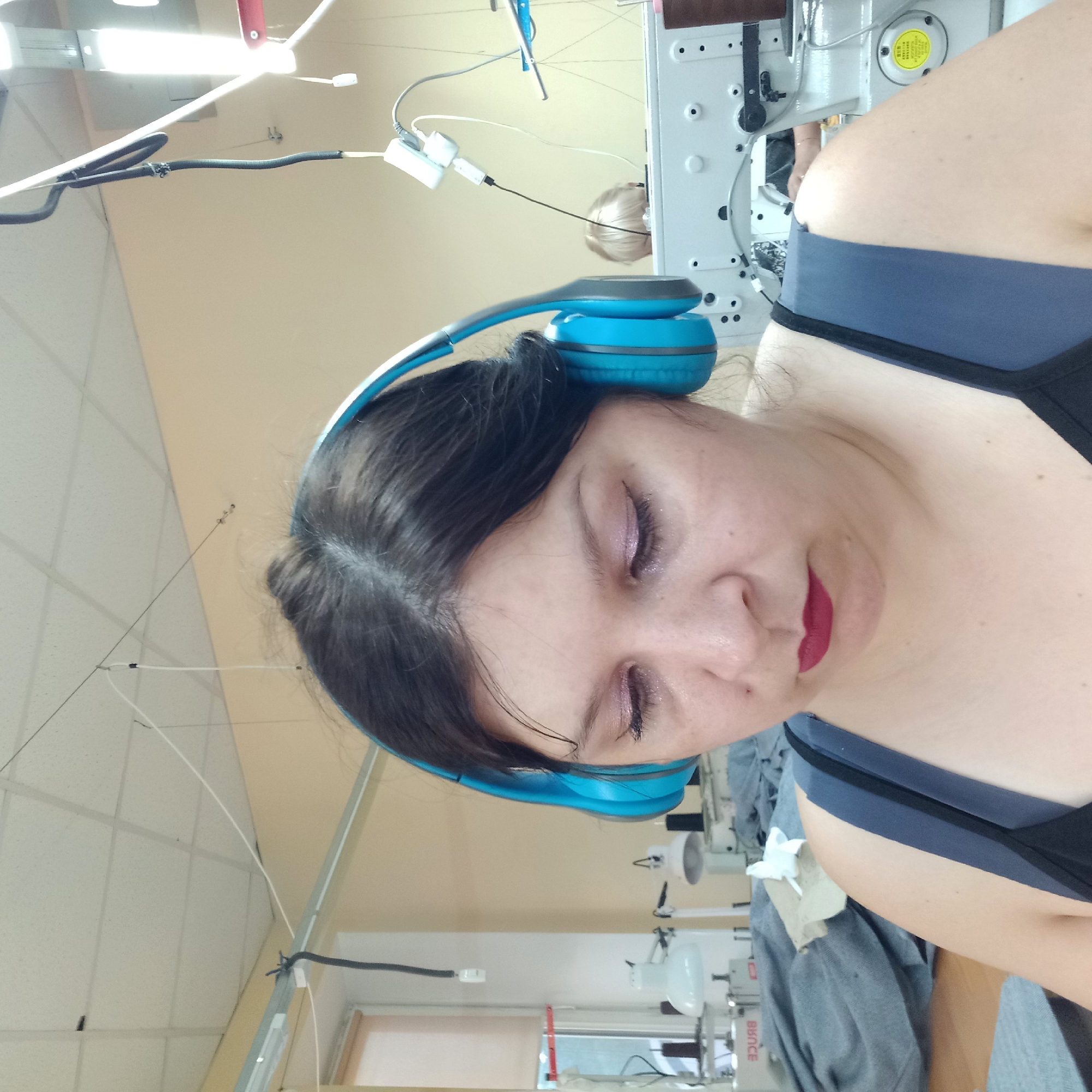 An indoor close-up shot shows a woman wearing blue headphones and looking down. Her dark hair is pulled back, and she has pink eyeshadow and dark red lipstick. She is wearing a dark tank top. In the background, industrial sewing machines and stacks of grey fabric are visible, suggesting a workshop or factory setting. The lighting is fluorescent. The overall scene appears to be in a commercial or industrial environment, possibly a garment factory.