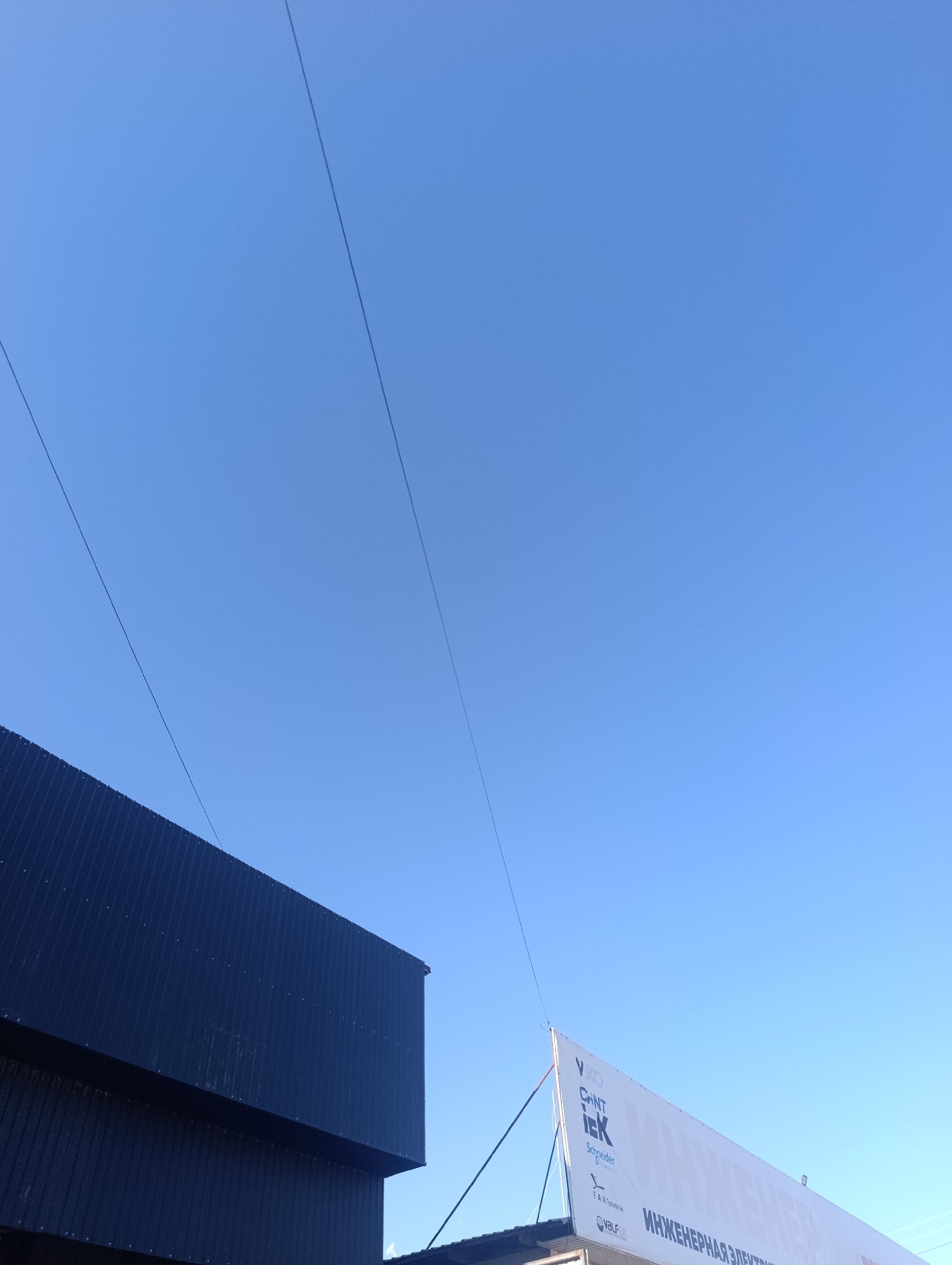 The image shows a clear blue sky above a modern dark-colored building on the left and a large white banner with text on the right. Three thin dark lines, likely wires or cables, stretch diagonally across the sky, originating from the top left and extending towards the bottom right. The building on the left has a corrugated facade. The banner on the right features several logos and text in Russian, including "ИНЖЕНЕРНАЯ ЭЛЕКТР" which translates to "ENGINEERING ELECTR". The scene is outdoors with no people visible. The image appears to be taken from a low angle looking upwards. The location is Bosteri, Kyrgyzstan.