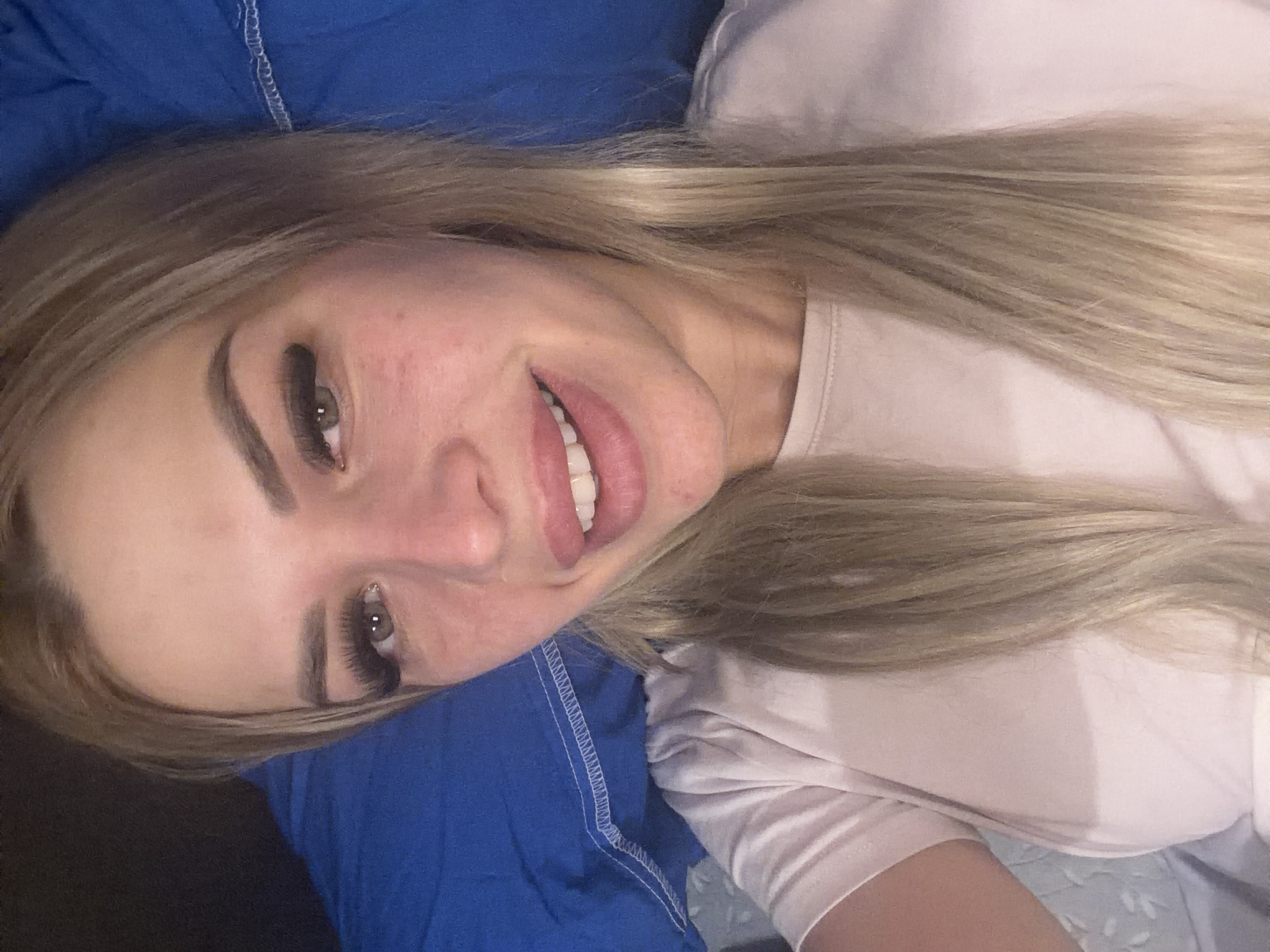 A close-up, high-angle, selfie-style shot shows a blonde woman lying down. She is wearing a light pink t-shirt and has on dramatic false eyelashes. Her mouth is open in a smile, revealing white teeth. The background is a blurry blue surface, possibly a pillow or blanket. There are no discernible landmarks or indications of a specific city or location.