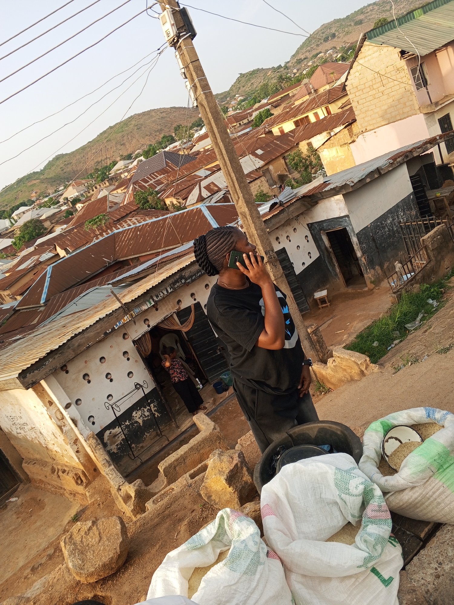 A woman in a black t-shirt and dark pants stands on a dirt street in Gwong, Nigeria, talking on a cell phone. The street is lined with weathered buildings with corrugated metal roofs and distressed walls. White sacks filled with grain sit on the ground near the woman. A utility pole stands near her. In the background are more buildings and a hillside.