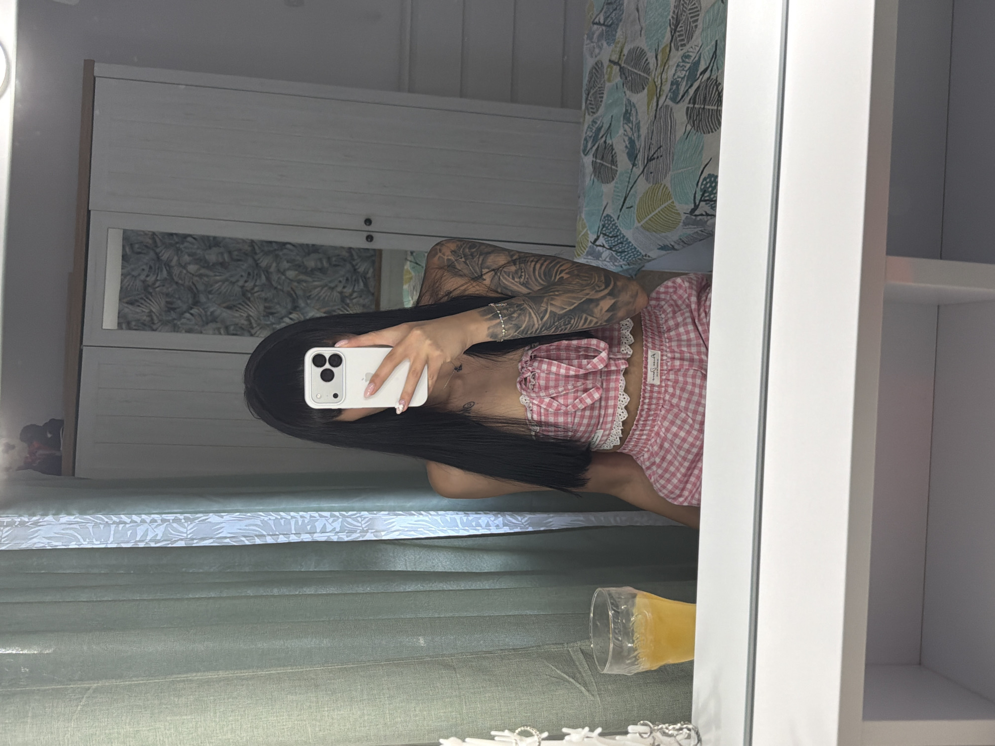 A young woman with long dark hair and bangs is captured in a mirror selfie taken in what appears to be a bedroom. She is wearing a pink gingham two-piece outfit, consisting of a bandeau top with lace trim and matching shorts. A glass of orange juice sits on a white vanity in front of her. Behind her, a closet with mirrored doors is visible, and a bed with a patterned duvet is to her right. The room is decorated with light green curtains. The image does not contain any specific visual cues that would allow for precise location identification within Plovdiv, Bulgaria.