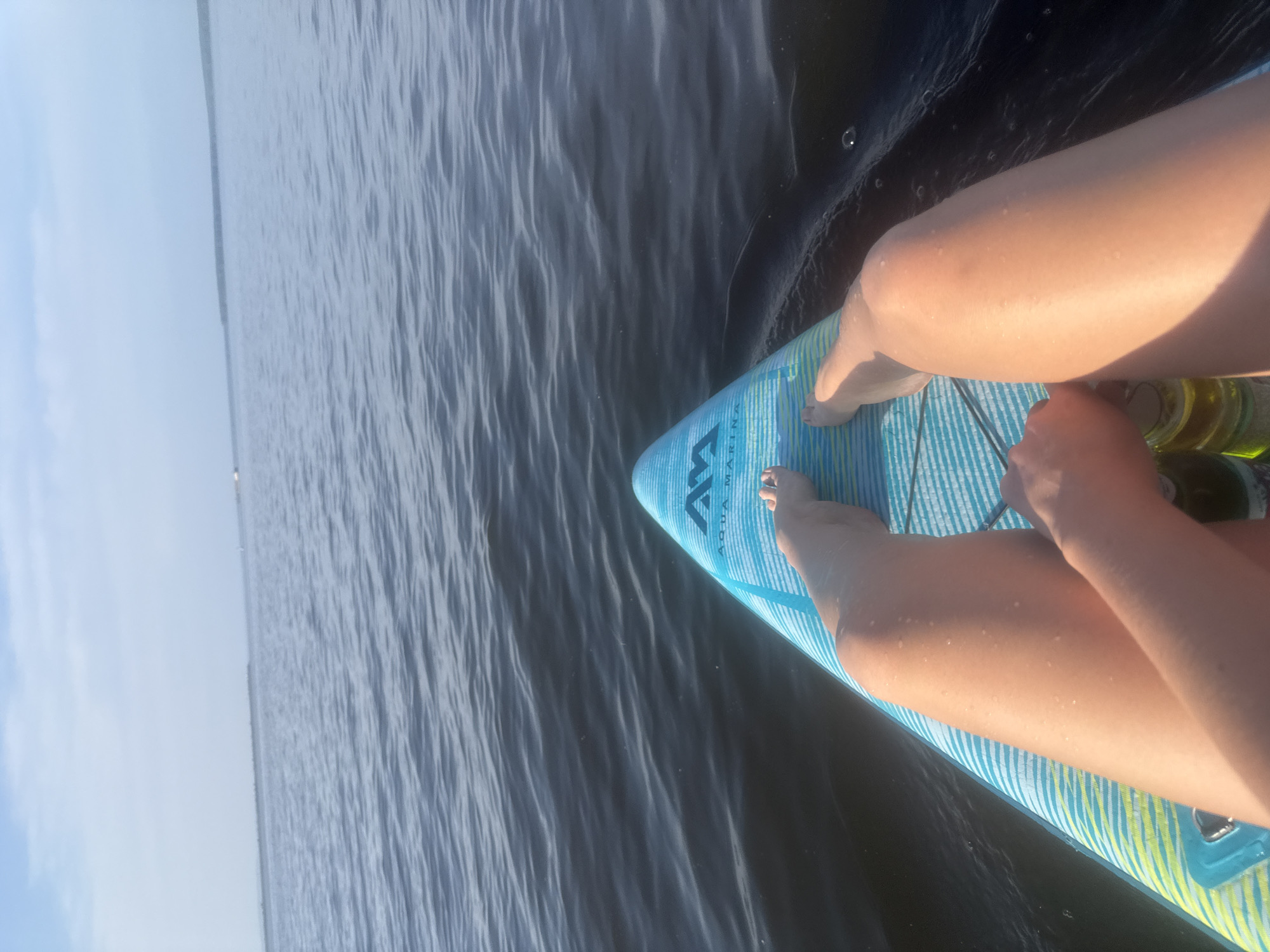 A person's legs are visible, resting on a blue and yellow striped paddleboard that floats on dark, rippling water. The person is holding two cans, one in each hand, with the right hand holding a can that is partially obscured by the left arm. The paddleboard has the "Aqua Marina" logo visible on its surface. The scene is set on a body of water, likely a river or lake, with a distant tree line on the horizon under a hazy sky.