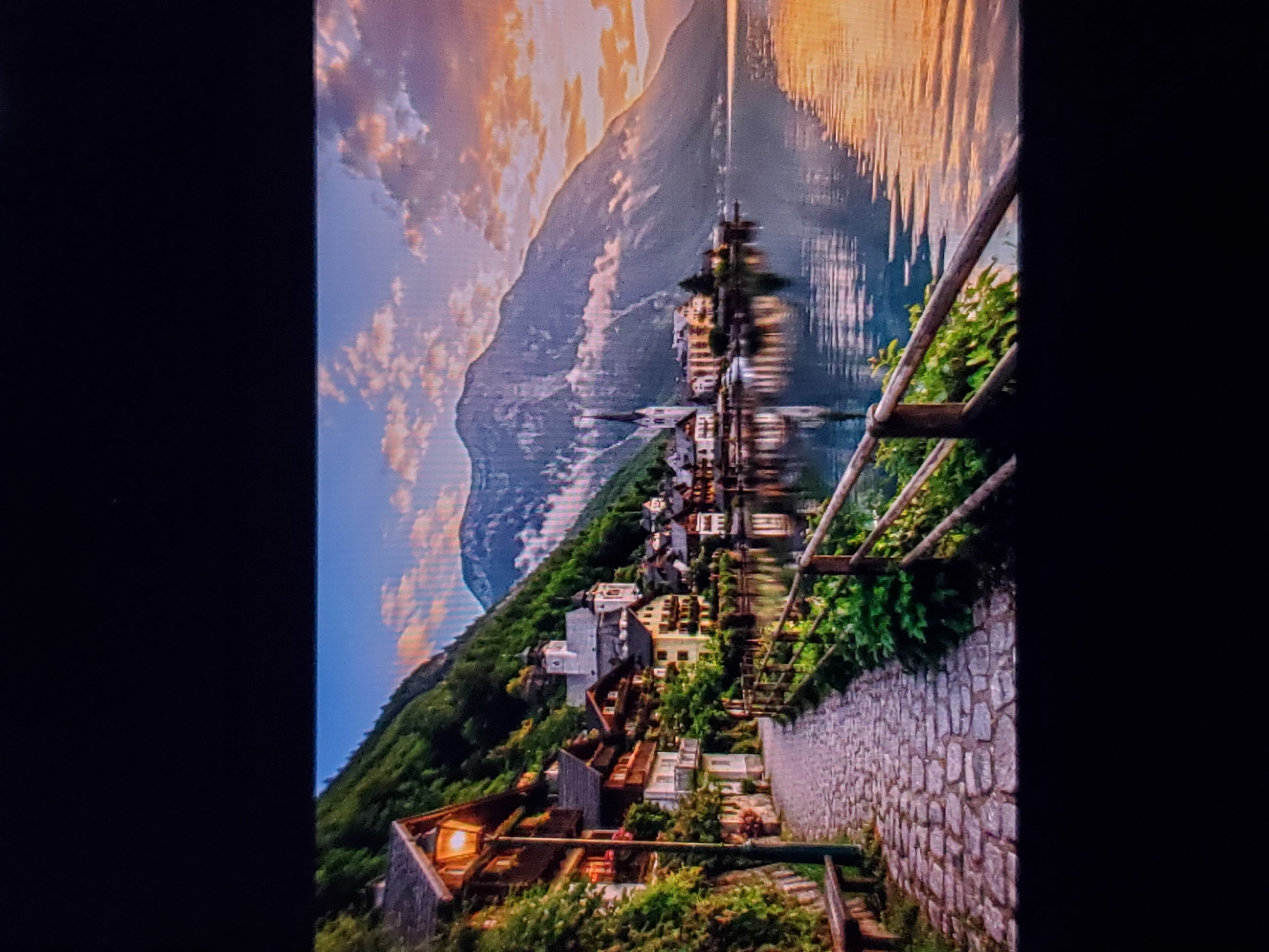 The image depicts a serene waterside village nestled at the base of a majestic mountain. A cobblestone pathway, illuminated by a vintage street lamp, winds alongside a row of traditional European-style houses. Lush greenery cascades down the hillside, framing the quaint buildings. Across the calm body of water, the mountain's imposing silhouette is softened by the warm hues of a sunset, casting a golden glow on the water's surface. A rustic wooden fence separates the path from the water, adding to the idyllic charm of the scene. The architecture suggests a European Alps setting, possibly a village in Austria like Hallstatt, known for its picturesque lakeside location and traditional houses.