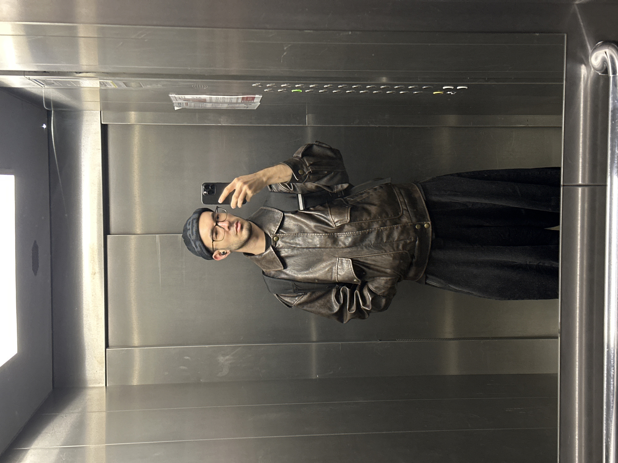 A slender male, with tan skin, is positioned in the middle of a stainless steel elevator. He has short brown hair and wears a gray beanie, black-rimmed glasses, a dark brown leather jacket, and dark clothing. The man is holding a smartphone in his left hand, taking a selfie. The elevator's interior is metallic and sterile, with horizontal panels and a metal handle visible on the right. A label is visible at the top of the elevator, though the specific details are obscured. The elevator car is brightly lit. The location is unidentifiable as Moscow.