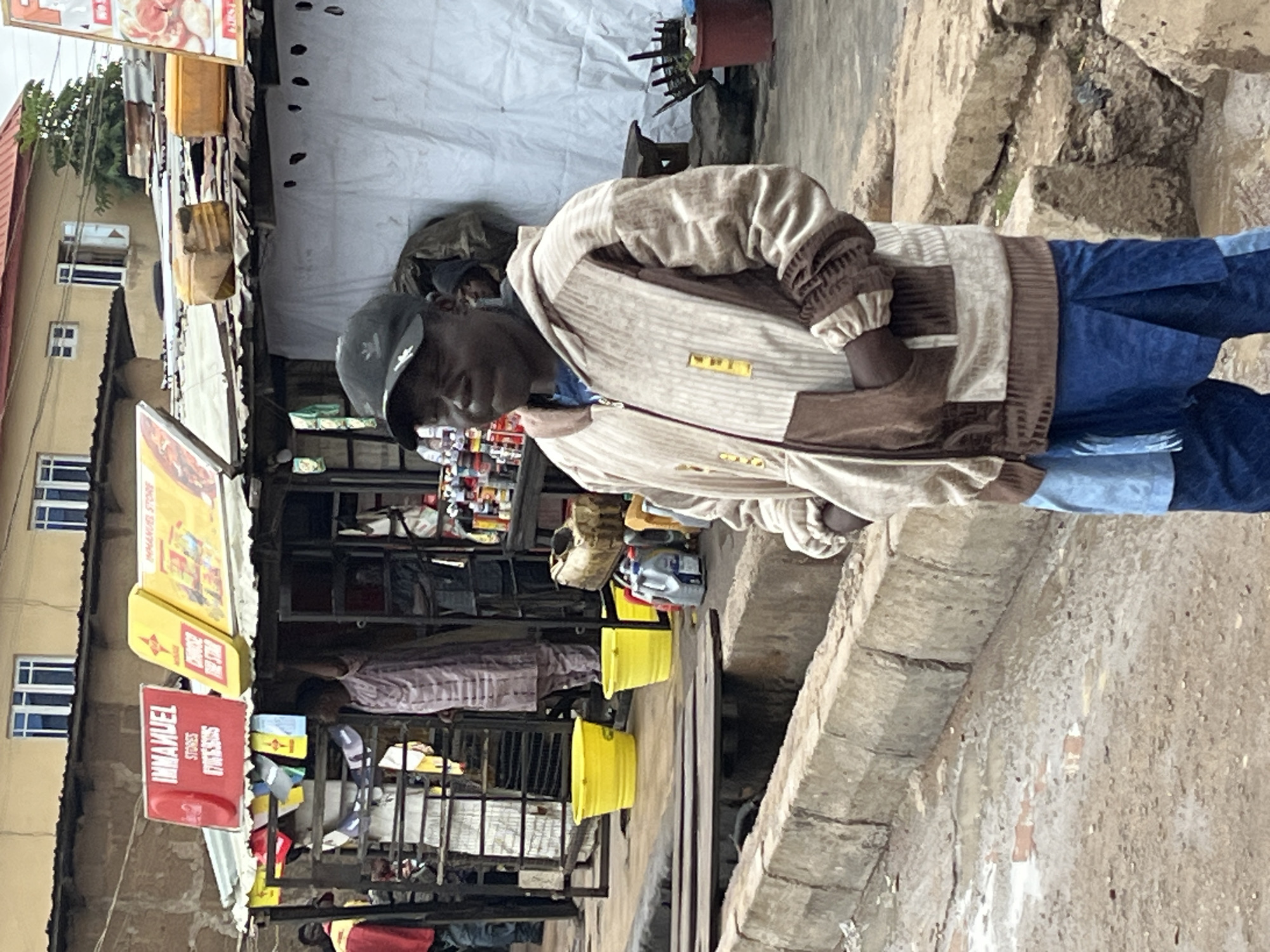A Black man wearing a black baseball cap, a tan and brown patterned jacket, and blue pants stands in front of a small market stall. The man is looking towards the left of the frame with his mouth slightly open. The market stall behind him has a corrugated metal roof and white sheeting on the sides. Several yellow containers, a red plastic bucket, and various bottles are visible on the ground near the stall. In the background, buildings with windows can be seen. The ground in front of the stall is wet and appears to be a street or walkway. The scene is outdoors in an urban environment.