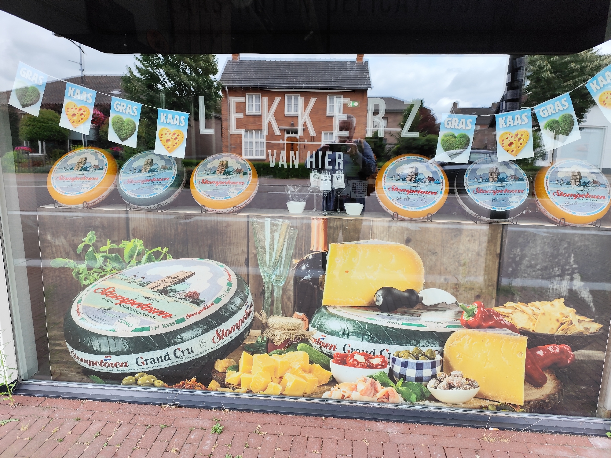 The image shows a Dutch cheese shop window in Schaijk, Netherlands. The window displays various cheeses, crackers, olives, and other food items. A string of banners with the words "GRAS" (grass) and "KAAS" (cheese) hangs across the top of the window. The text "LEKKER VAN HIER" (delicious from here) is visible on the window, referring to local produce. A man is reflected in the window, taking a photo. The shop appears to be located in a residential area with houses and trees visible in the background. The weather is overcast.