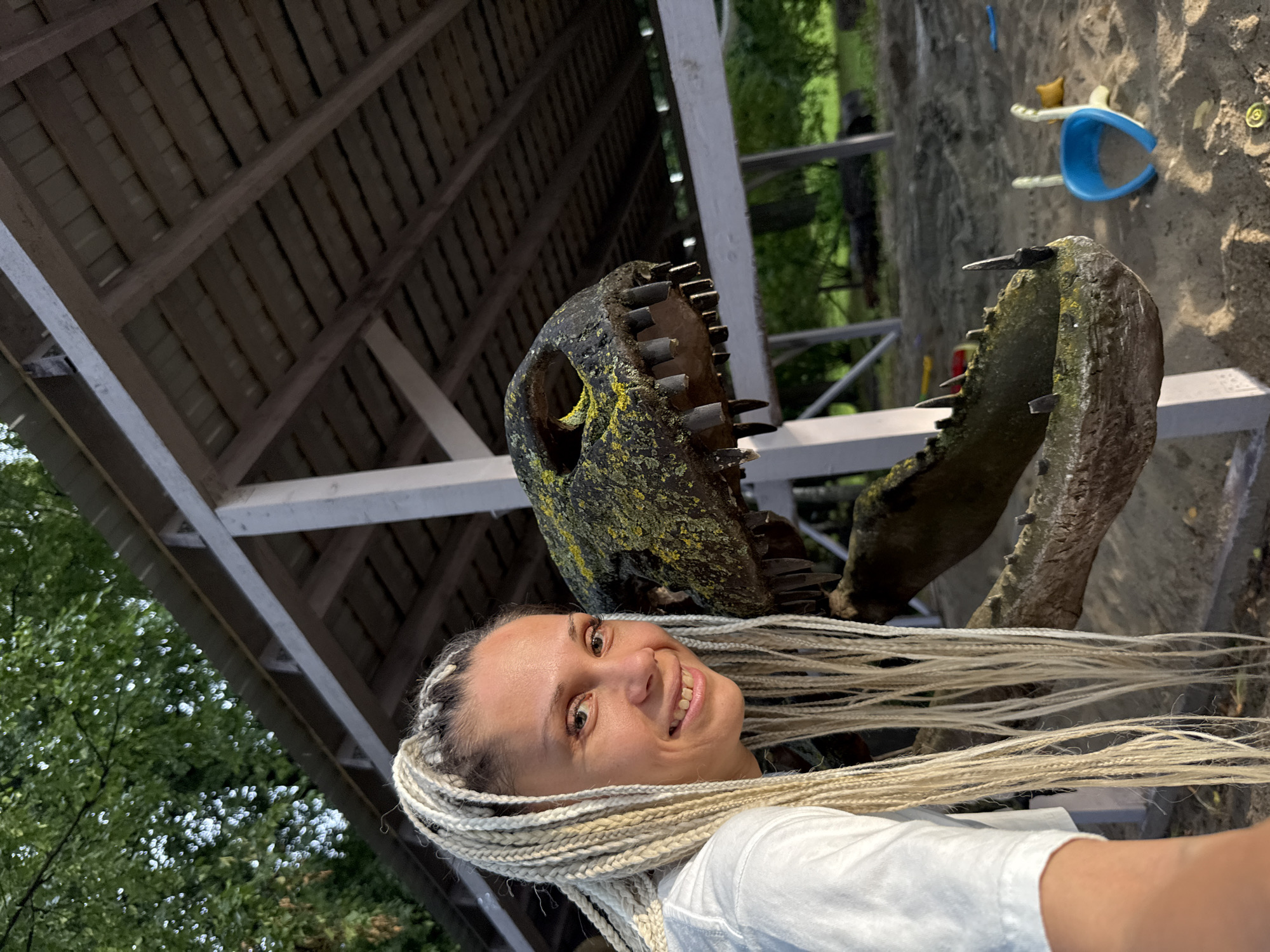 The image shows a woman with blonde braided hair wearing a white t-shirt, smiling at the camera. She is positioned to the left of a dinosaur skull model, which is weathered and has visible teeth. The dinosaur model appears to be part of a playground or outdoor exhibit, as indicated by the sand-covered ground and some scattered toys in the background, including a blue and yellow toy. Behind them, there is a wooden structure with a covered roof and supporting beams. The environment suggests an outdoor recreational area. The image appears to be taken in Rusanovo, Russia.