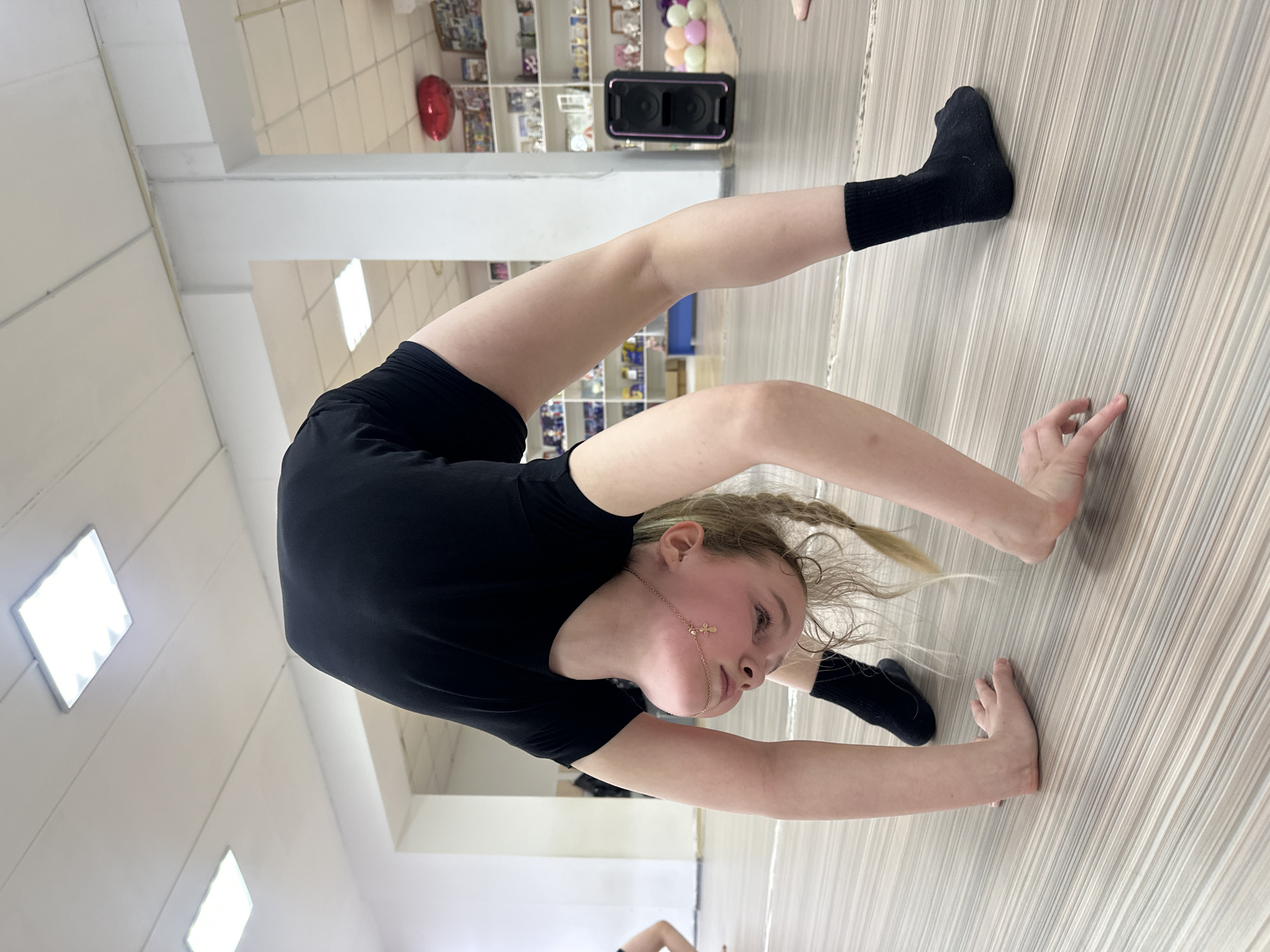 A young girl in a black leotard and black socks is performing a backbend on a light-colored wood floor. She is using her hands for balance. Her hair is blonde and braided. The location appears to be a dance studio. A small speaker is visible in the background, along with shelves containing various items, including a red balloon. While precise location data requires explicit metadata or verifiable clues within the image, the presence of unique cultural items may assist in future localization to the region of Votkinsk, Russia.