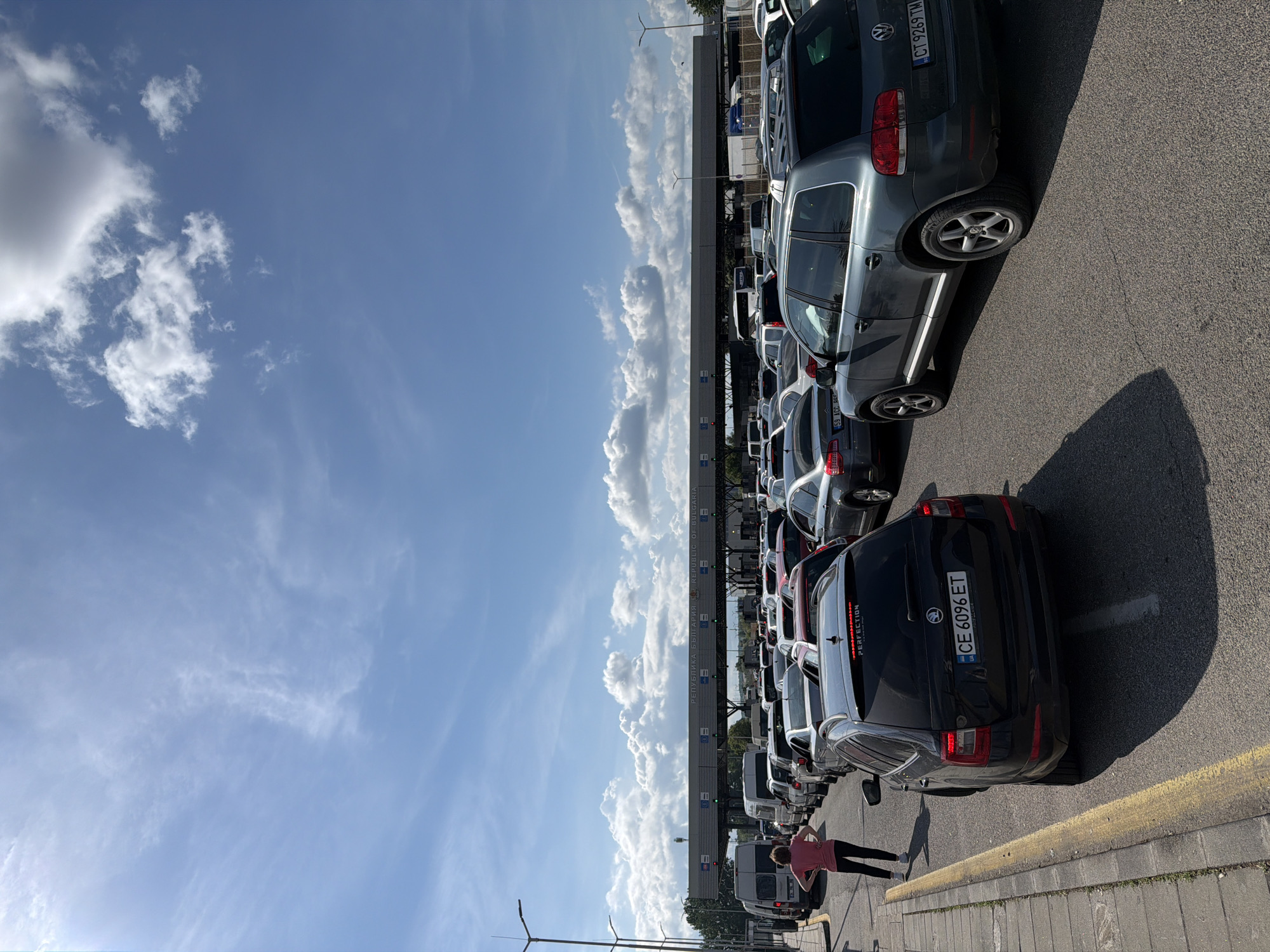 The image shows a line of cars waiting at the Kapitan Andreevo border crossing between Bulgaria and Turkey. The camera captures the scene from an elevated point along the roadside, looking towards the border checkpoint. The sky takes up the left portion of the image and is blue with puffy white clouds. A border control booth with signage in both Bulgarian and English is visible, reading, "REPUBLIKA BULGARIA - REPUBLIC OF BULGARIA". The main subject is a line of stationary vehicles waiting to cross the border, mostly passenger cars, and a van. A pedestrian is visible walking alongside the parked cars. The ground is paved. Overhead power lines are also present in the lower-left corner. A yellow line is painted along the pavement.