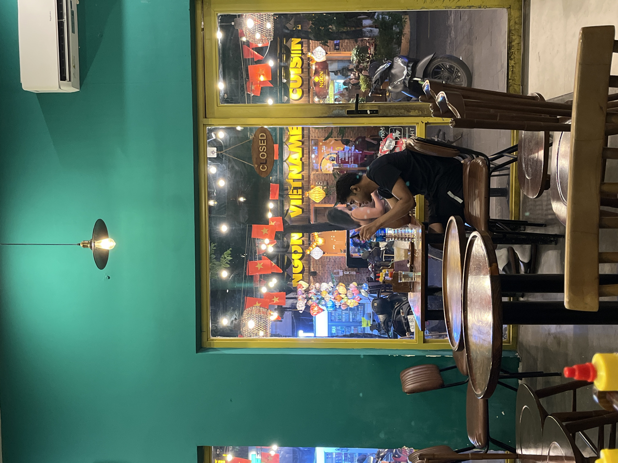 The photo was taken indoors, with an open window onto a bustling street in what appears to be Đà Nẵng, Vietnam. The interior is painted a vibrant teal. A couple is standing at a table, interacting with their phones in a restaurant. Looking through the window, a Vietnamese restaurant is visible with a sign reading "VIETNAM" and another indicating "NGON CUISINE." Lanterns and flags are also adorning the street scene. Motorbikes are parked along the street. Chairs are stacked up and table tops are visible in the foreground, hinting that the restaurant may be closed or preparing to open.