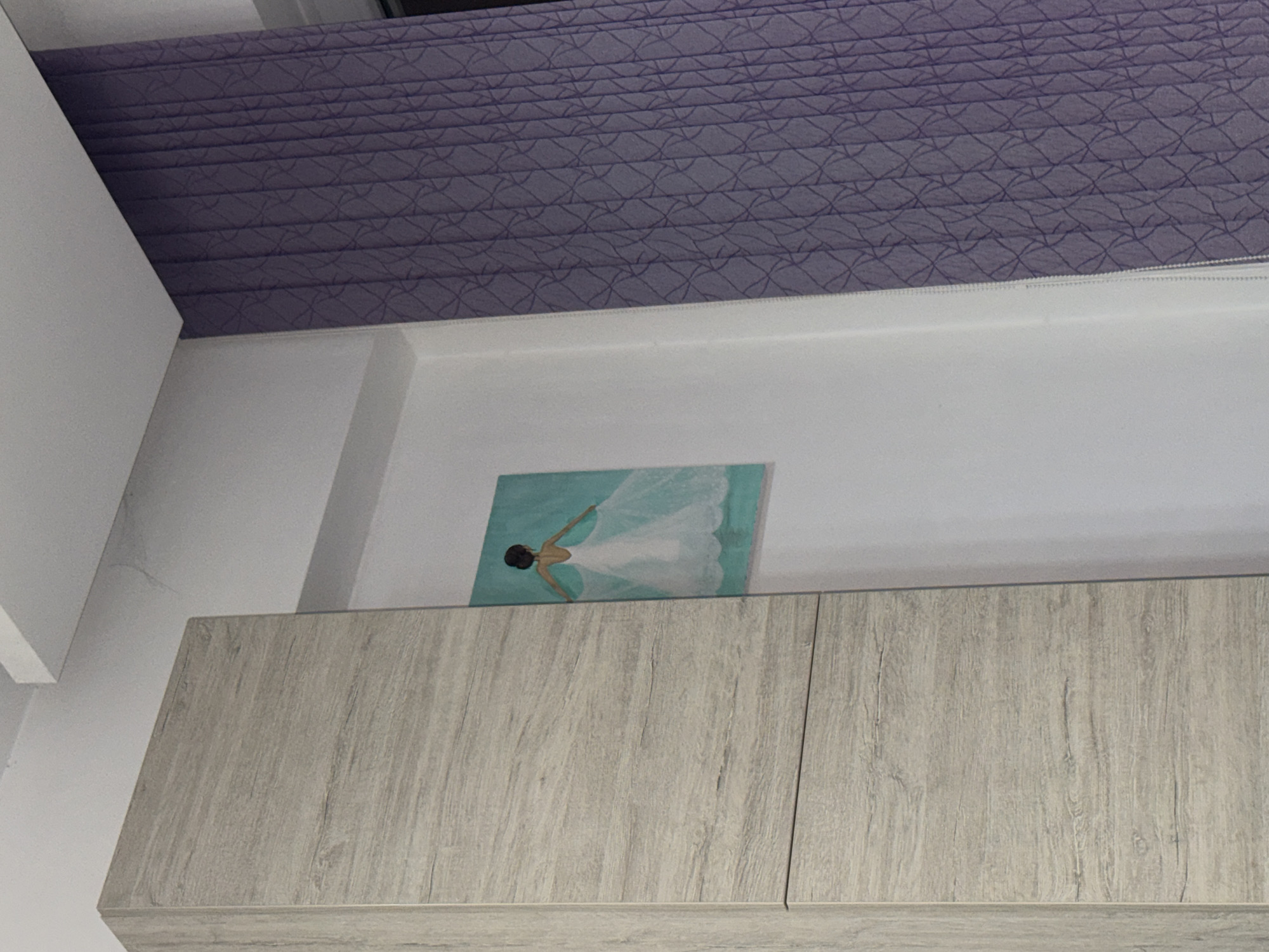 A low-angle shot shows a section of a room, focusing on a set of cabinets with a light wood-grain finish. On top of the cabinets rests a small, framed painting with an aquamarine background, depicting a figure in a white dress. Above the painting and cabinets is a white wall with a purple, patterned roller blind partially drawn down. The scene takes place indoors. Without additional information, the location in Plovdiv, Bulgaria cannot be determined.