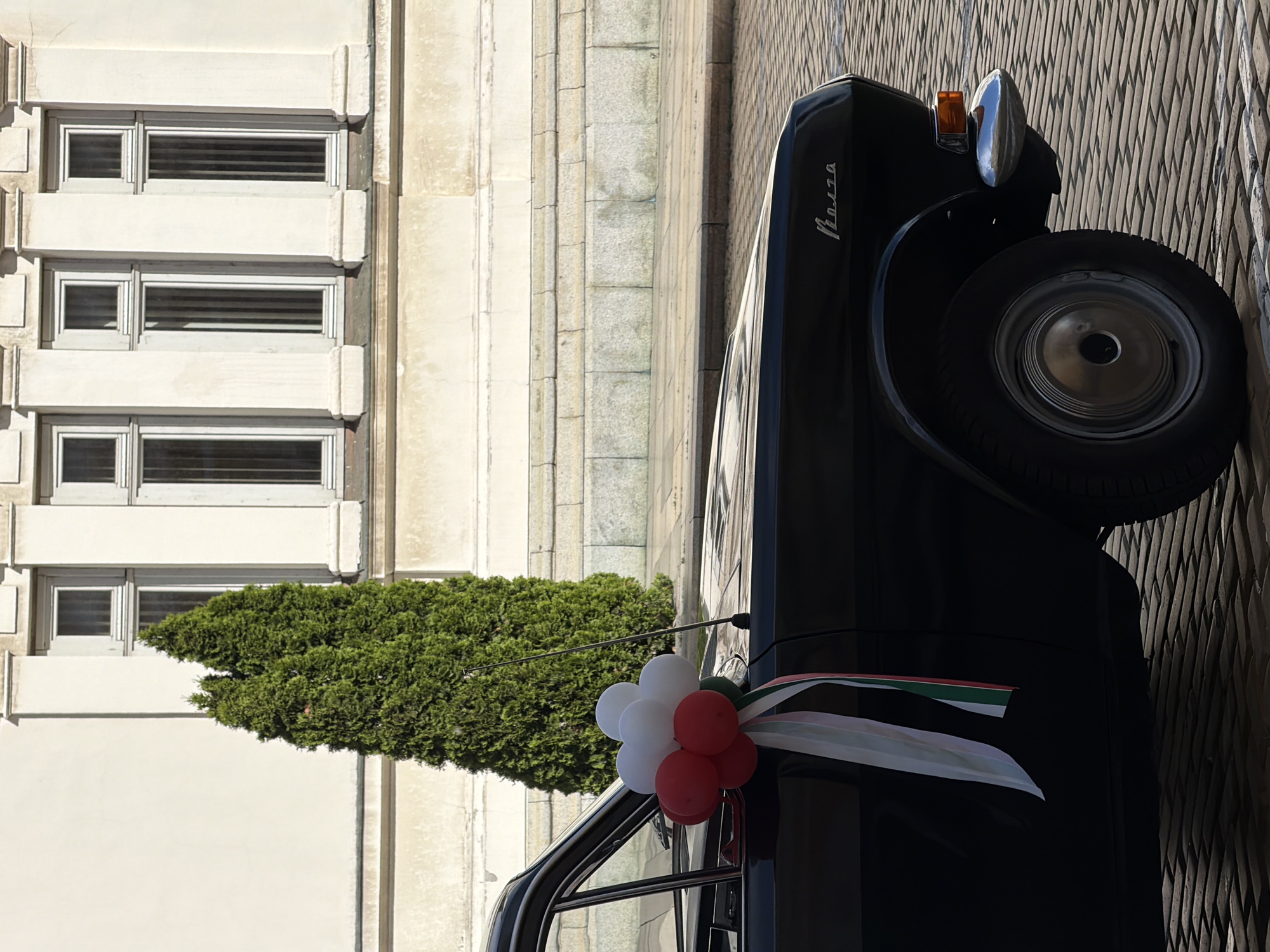 A black Moskvich Aleko is parked next to a light-colored building with windows and a trimmed tree. The car is decorated with red and white balloons and an Italian flag ribbon. The location is Sofia, Bulgaria.