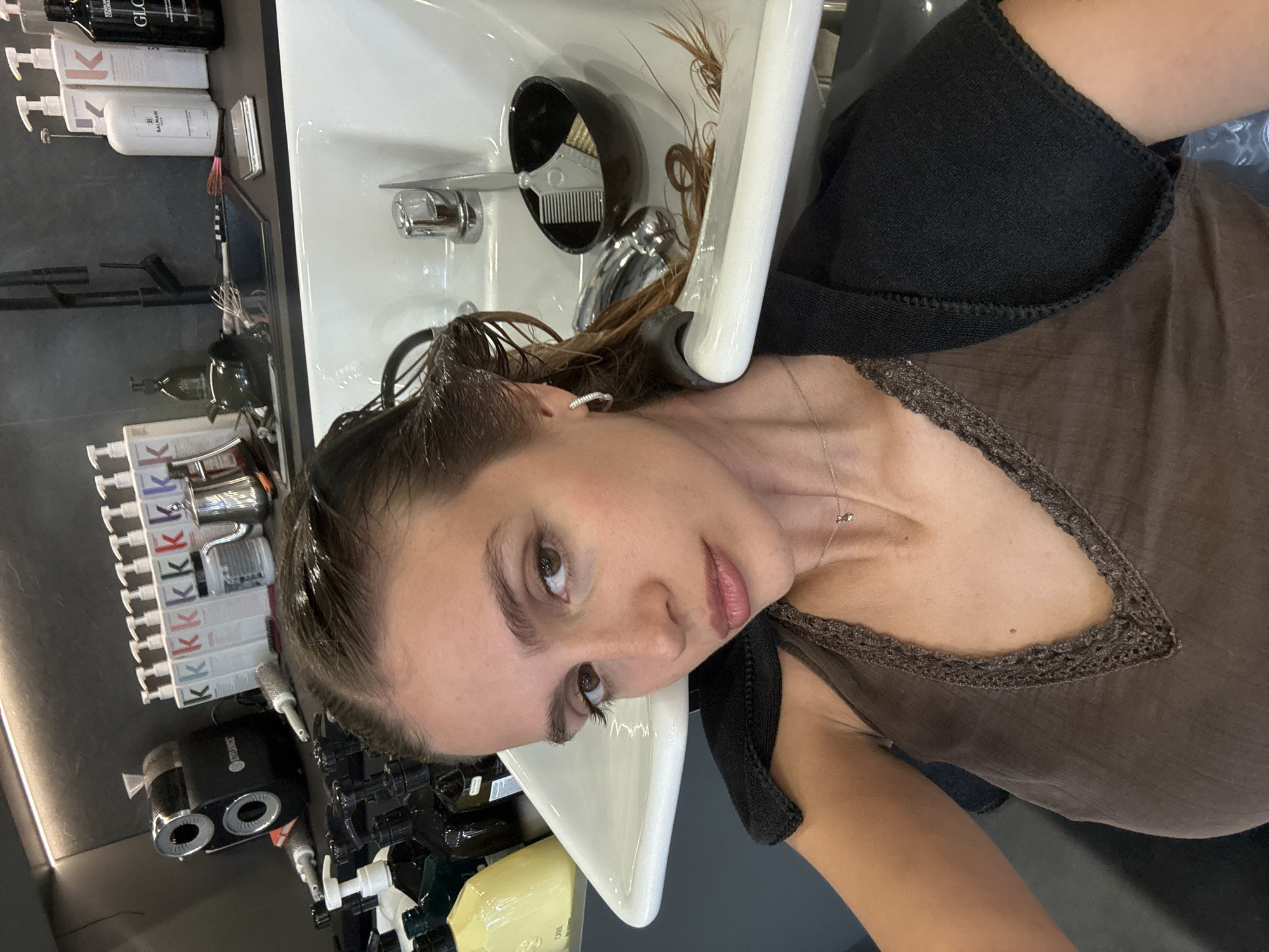 The image shows a woman with light brown hair having her hair washed at a salon. Her head is tilted back over a white porcelain sink with a black comb attached to the side. The sink has a chrome faucet. She is wearing a brown tank top with a lace trim and a black towel around her neck. There are several bottles of hair products lined up on a shelf to the left, including bottles with the letter "K" on them. The environment appears to be a professional salon, with tiled walls and various hair care products and equipment visible. The location is likely Tbilisi, Georgia.