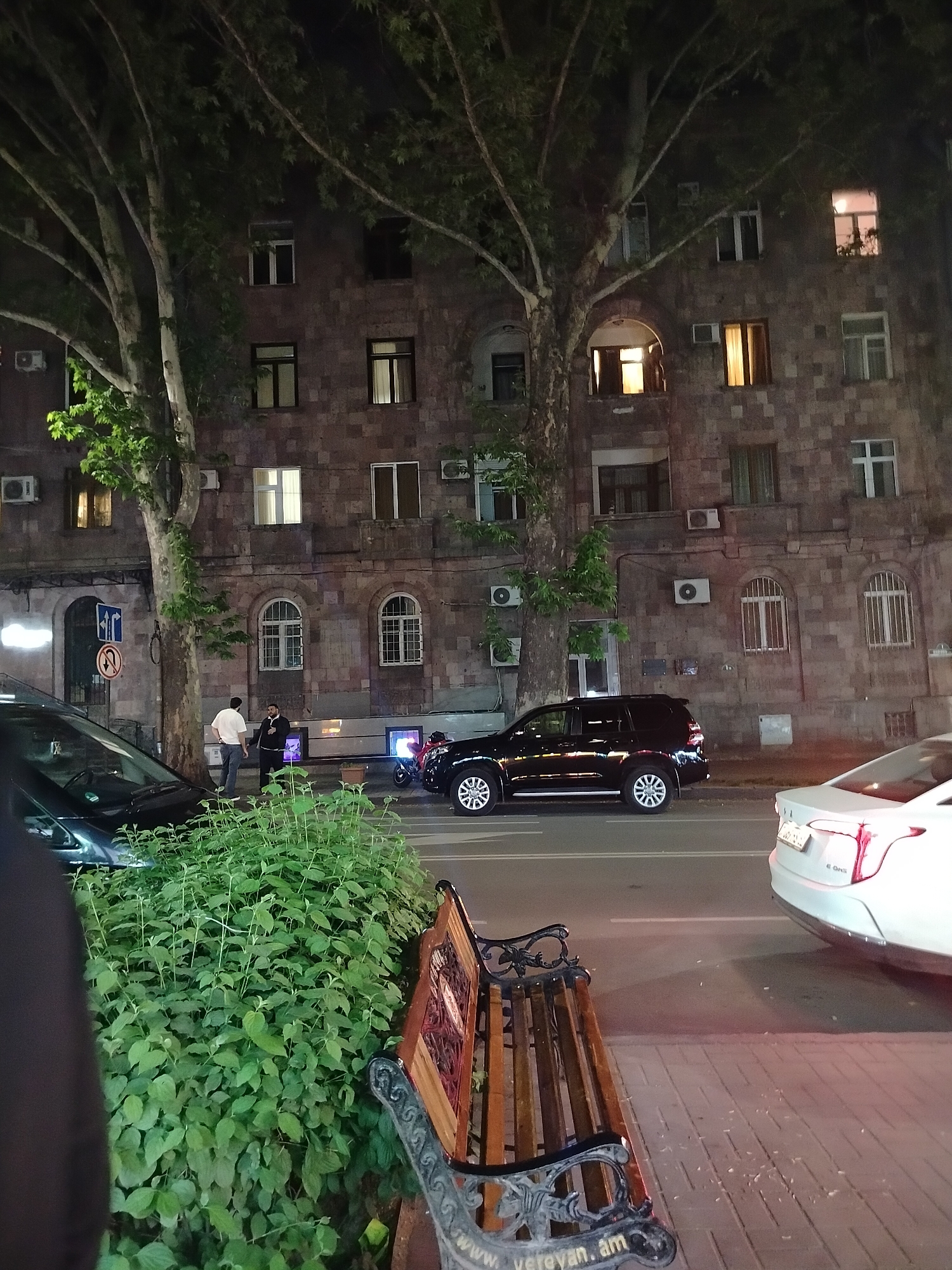 At night, in Yerevan, Armenia, a street scene unfolds in front of a multi-story stone building with many windows, some illuminated. Two men are standing on the sidewalk near a black car parked in front of the building. Several other cars are parked along the street. A green bush and a park bench with "Yerevan.am" inscribed on it are visible in the foreground. Large trees line the street, partially obscuring the building and street lighting. Air conditioning units are attached to the building's exterior walls.