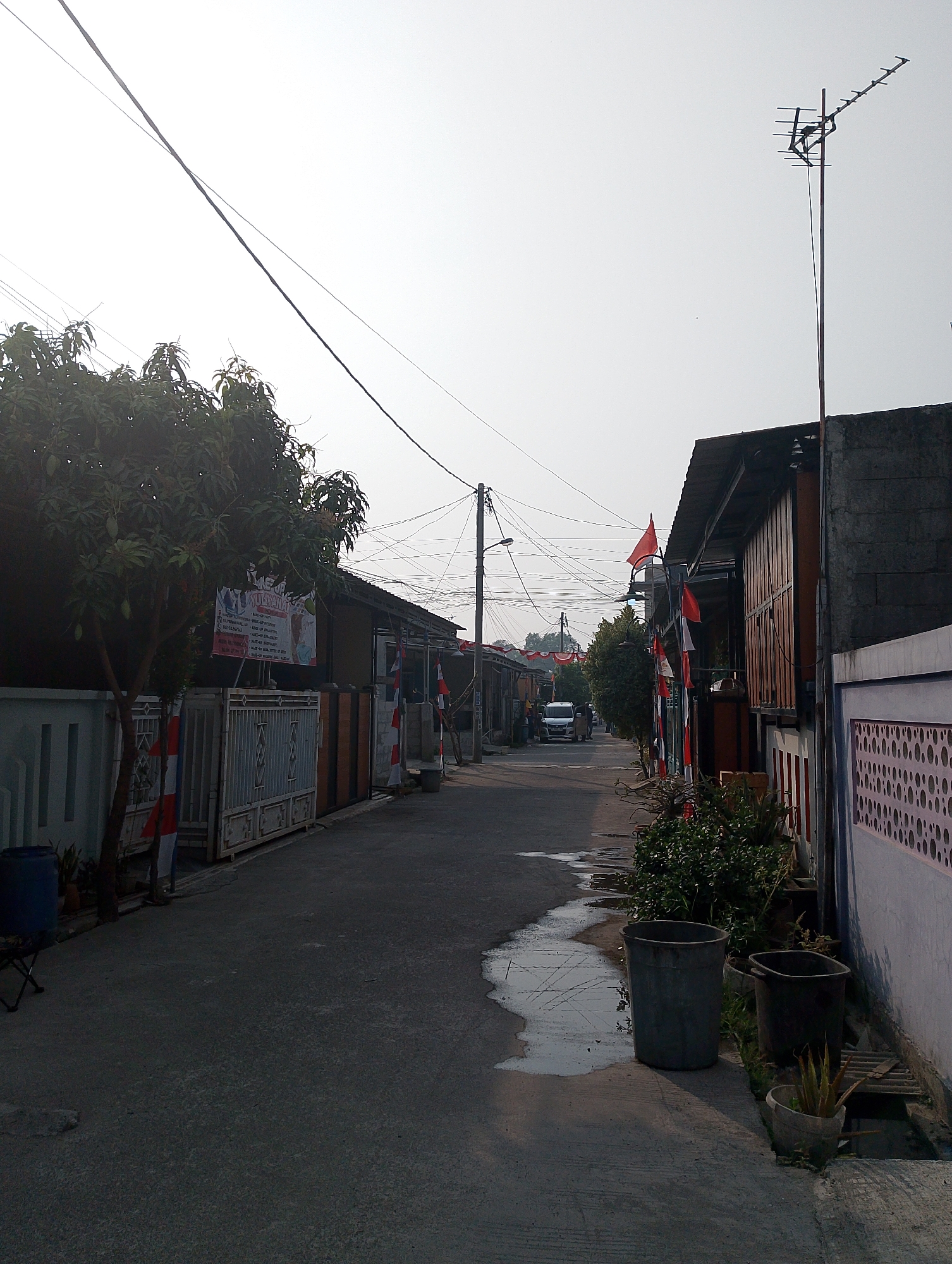 A residential street in Karawang, Indonesia, is depicted on a bright, clear day. The street is paved and appears to be a quiet, local thoroughfare. On the left side of the street, a tree with green foliage is prominently featured, with a white fence and a blue barrel visible nearby. A banner is attached to the tree, displaying text in Indonesian. Further down the street, on both sides, are houses with attached fences and gates. Several Indonesian flags are displayed, attached to poles and buildings, suggesting a national holiday or celebration. The houses are of varied architectural styles, with some featuring wooden elements and corrugated metal roofs. Overhead, a complex network of electrical wires crisscrosses the sky. In the distance, a white van is visible, along with a few pedestrians. On the right side of the street, more houses are visible, with one wall adorned with a repeating decorative pattern. Several large trash bins are placed along the sidewalk, along with potted plants. A small puddle of water is present on the road, reflecting the overcast sky. An old television antenna stands tall above one of the houses on the right. The scene suggests a typical day in a residential neighborhood in Indonesia, with elements of local culture and daily life clearly visible.