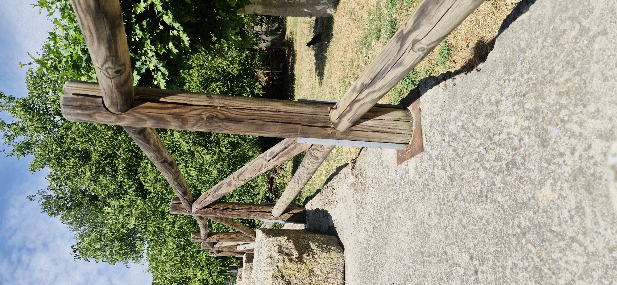 An outdoor, low-angle shot captures a rustic wooden fence with rough-hewn posts and rails. The fence runs diagonally from the lower left to the upper right of the frame. To the left of the fence, a large, light-colored rock is visible, partially obscuring the concrete path in the foreground. In the background, lush green trees form a dense canopy, suggesting a natural or park-like setting. The ground beyond the fence is covered in dry grass, with a small black cat silhouetted against the sunlight in the middle distance. The sky above is a clear blue with scattered white clouds.