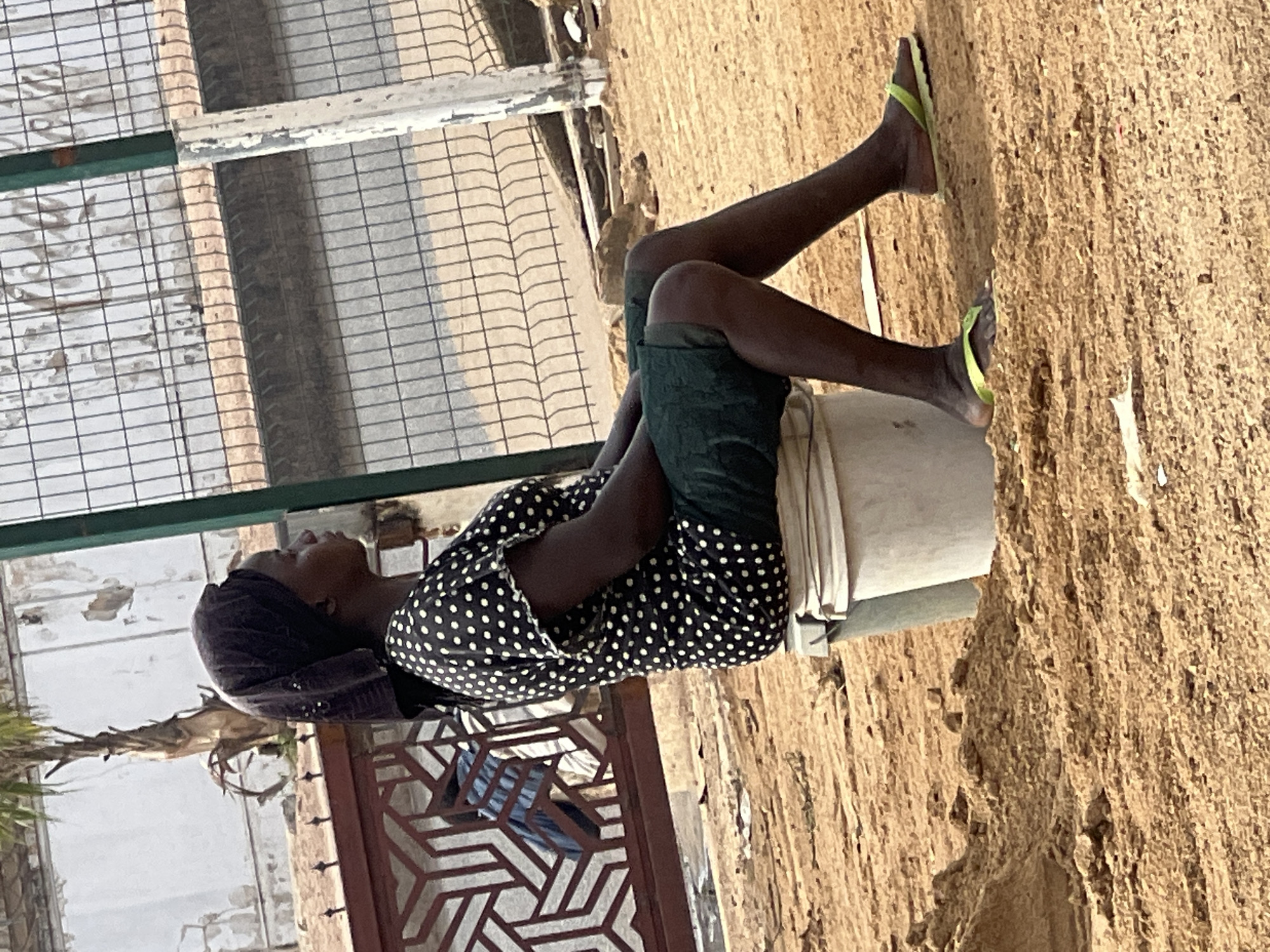 A woman wearing a black and white polka dot shirt, dark green shorts, and a dark head covering is sitting on a white bucket. She is wearing lime green flip-flops. The woman is sitting near a building with a metal gate and wire fence. The ground is dirt. The location is Barakin Lerawi, Nigeria.