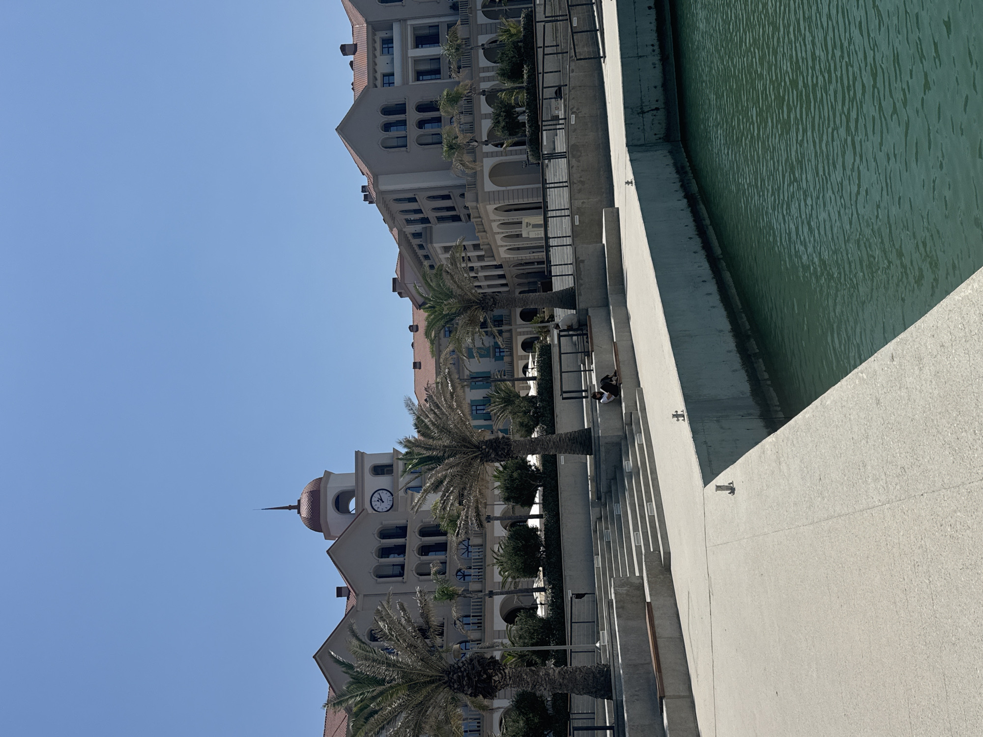 This is a wide shot of a waterfront promenade in Sabunchu Raion, Azerbaijan, under a clear blue sky. In the background, a multi-story building with arched windows and red-tiled roofs dominates the scene, featuring a prominent clock tower. Palm trees are scattered throughout the area, and a person is seated on a concrete ledge near the water. The promenade itself is made of concrete, with tiered steps leading up from the water's edge. The water is a dark teal color, appearing calm.