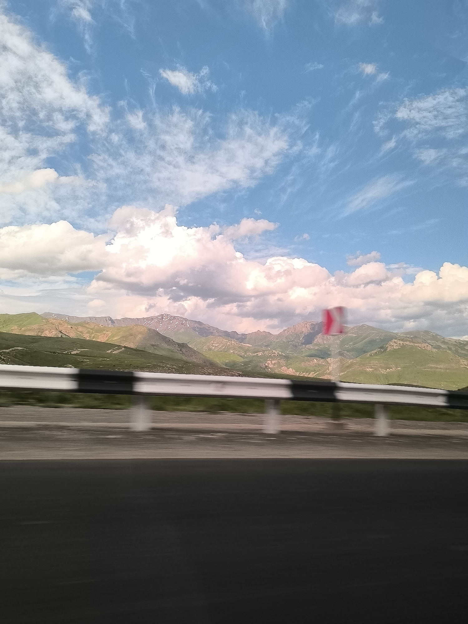 The image captures a vibrant mountainous landscape as seen from a moving vehicle, likely on a highway in Yelpin, Armenia. The scene is characterized by distinct horizontal layers.
In the immediate foreground, a dark asphalt road surface occupies the bottom third of the frame, exhibiting significant motion blur, which strongly suggests the photo was taken from a vehicle in transit. Just above the road runs a white and black striped metal guardrail, also blurred, reinforcing the sense of movement.
The midground features an extensive range of mountains and hills. These formations are largely covered in rich green vegetation, indicating a lush environment. Some higher, more distant peaks show lighter patches, possibly of exposed rock or lingering snow. The mountains are illuminated by bright daylight, casting subtle shadows and highlighting contours. A blurry red and white traffic sign, possibly a warning for a curve, is partially visible on the right side of the midground.
The sky dominates the upper portion of the image, displaying a clear, bright blue color. It is dotted with scattered white clouds of varying types. Fluffy cumulus clouds are prominent, some exhibiting a slight pinkish or purplish tint, possibly due to atmospheric conditions or the angle of the sunlight. Wispy, thin cirrus clouds are also visible, adding texture to the expansive sky.
The image conveys a sense of travel through a grand natural setting during a clear, sunny day. No people or discernible text are visible.