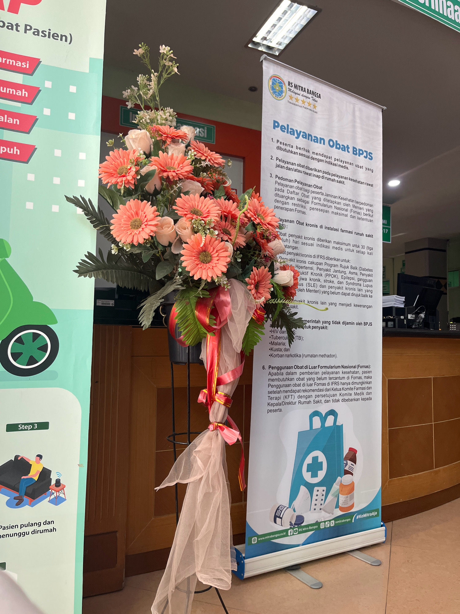 The image depicts a floral arrangement in front of a vertical banner advertising services at RS Mitra Bangsa in Kecamatan Pati, Indonesia. The banner, predominantly white with blue and green accents, details "Pelayanan Obat BPJS" (BPJS Drug Services), outlining patient rights, drug dispensing procedures, and coverage for chronic conditions. It features text in Indonesian, a logo for RS Mitra Bangsa, and social media icons. The floral arrangement consists of pink and peach gerbera daisies, white roses, and green foliage, tied with sheer pink and red ribbons. To the left of the banner, a vertical sign with a green background and white polka dots illustrates a scooter and lists services like "Farmasi" (Pharmacy), "Rumah" (Home), "Jalan" (Street), and "Puskesmas" (Community Health Center). An illustration of a person relaxing on a sofa is visible on this green sign, labeled "Step 3." The background shows a wooden counter and what appears to be a reception area within a medical facility.
