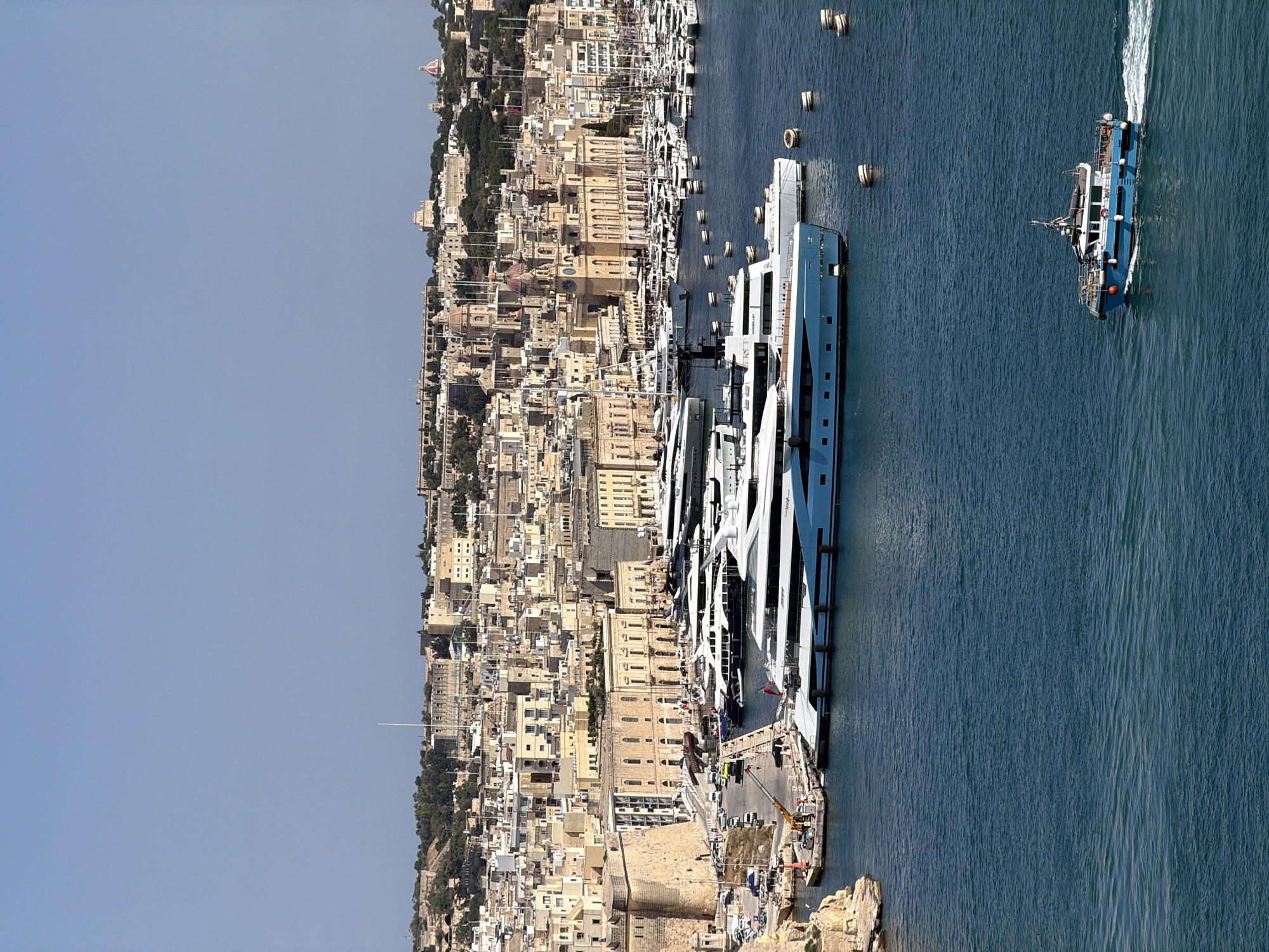 This aerial photograph captures a harbor scene in Valletta, Malta. The main subjects are the docked superyachts and a working boat in the water. The scene is a bustling port with a cityscape of beige buildings rising behind the waterfront. The time of day appears to be midday, indicated by the bright sunlight and clear blue sky. The weather is fair.
The large, modern superyachts are docked along the waterfront, showcasing sleek designs and white exteriors. A smaller blue and white working boat is actively moving through the water, leaving a wake. In the background, the historic architecture of Valletta is visible, with its characteristic stone buildings and red-domed churches. Cranes and construction equipment suggest ongoing activity at the port. The water is a deep blue, reflecting the sky.
There are no discernible people in this image, though there might be crew on the boats or workers on the docks, they are too small to be identified. No text is clearly visible on any of the objects or structures in the image. The overall impression is one of a vibrant maritime hub, combining modern luxury with historical context.