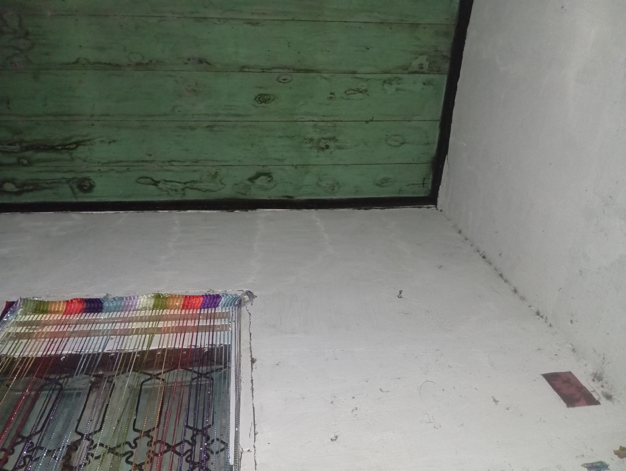 In the city of Rejosari, Indonesia, this image depicts a corner of an interior room. The upper portion of the image is dominated by a dark green wooden ceiling made of horizontal planks, showing signs of wear and discoloration. Below the ceiling, a white-plastered wall extends downwards, meeting at a sharp angle to form the corner of the room. A dark border runs along the junction of the ceiling and the wall.
On the lower left side of the white wall, a decorative beaded curtain is visible, hanging from the top of what appears to be a window or doorway. The curtain is colorful, with bands of various hues like purple, blue, green, yellow, orange, and red at the top, transitioning into strings of metallic beads of different colors, including silver and dark blues and purples. The curtain hangs over a dark grid-like structure, possibly a window grill.
The white wall itself is textured and shows some imperfections, including faint lines, small dark spots, and a small piece of red material affixed to it on the right side. A small, L-shaped object, possibly a nail or hook, is also present on the wall in the upper right quadrant. The lighting in the room appears to be artificial and casts subtle shadows, highlighting the textures of the walls and ceiling.