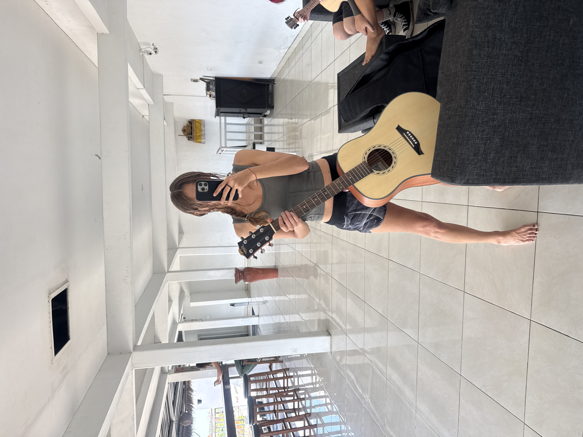 The image shows a person holding a guitar in what appears to be an open-air structure with white tiled flooring and white support beams. The person is taking a selfie using a smartphone; their face is partially obscured by the phone. They are wearing a gray tank top and patterned shorts. There is another guitar in the background, along with what looks like a guitar case and another individual. The environment appears to be a building with a view of the outside, where one can see green vegetation and possibly a body of water. The image appears to be taken in a structure that has a partial roof and a view to the outside. The location is likely in North Kuta, Indonesia.